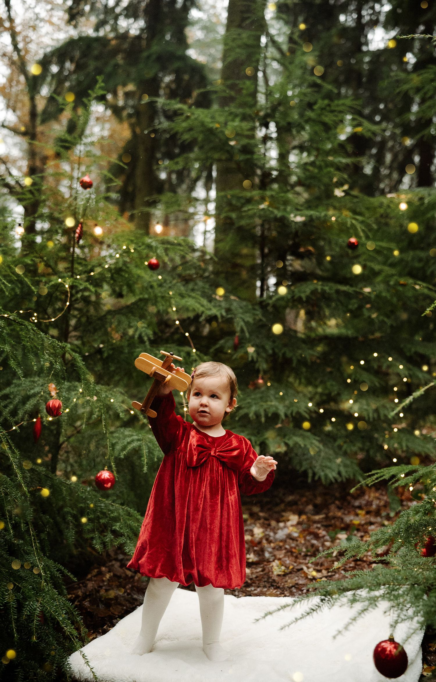 Toddler in red velvet dress holding wooden toy beside decorated Christmas trees with gold and red ornaments outdoors.