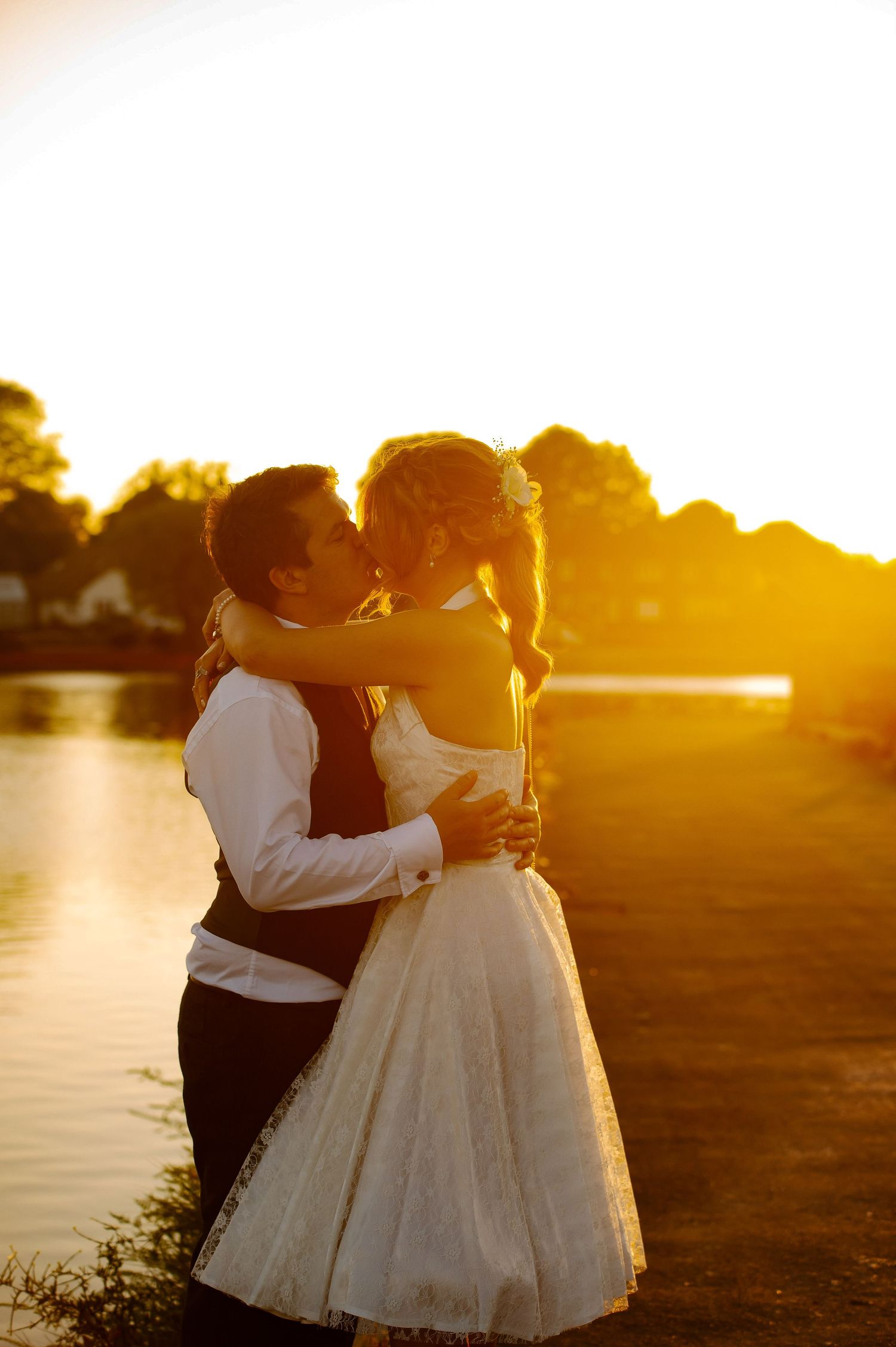 A couple in wedding attire embraces by a lake during golden hour sunset, creating a romantic silhouette.