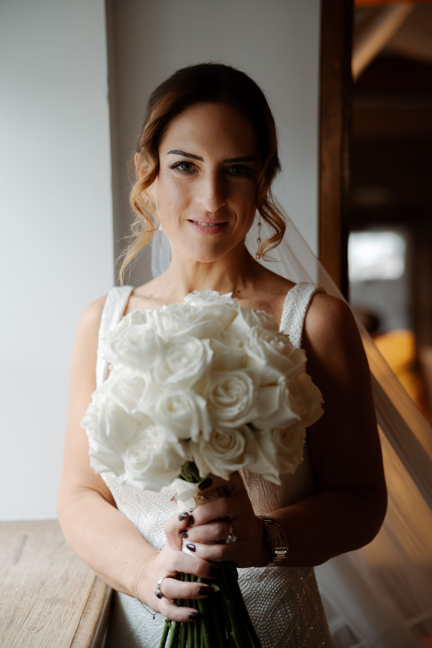 Bride in white gown holding elegant bouquet of white roses, wearing veil and jewelry, posing indoors.