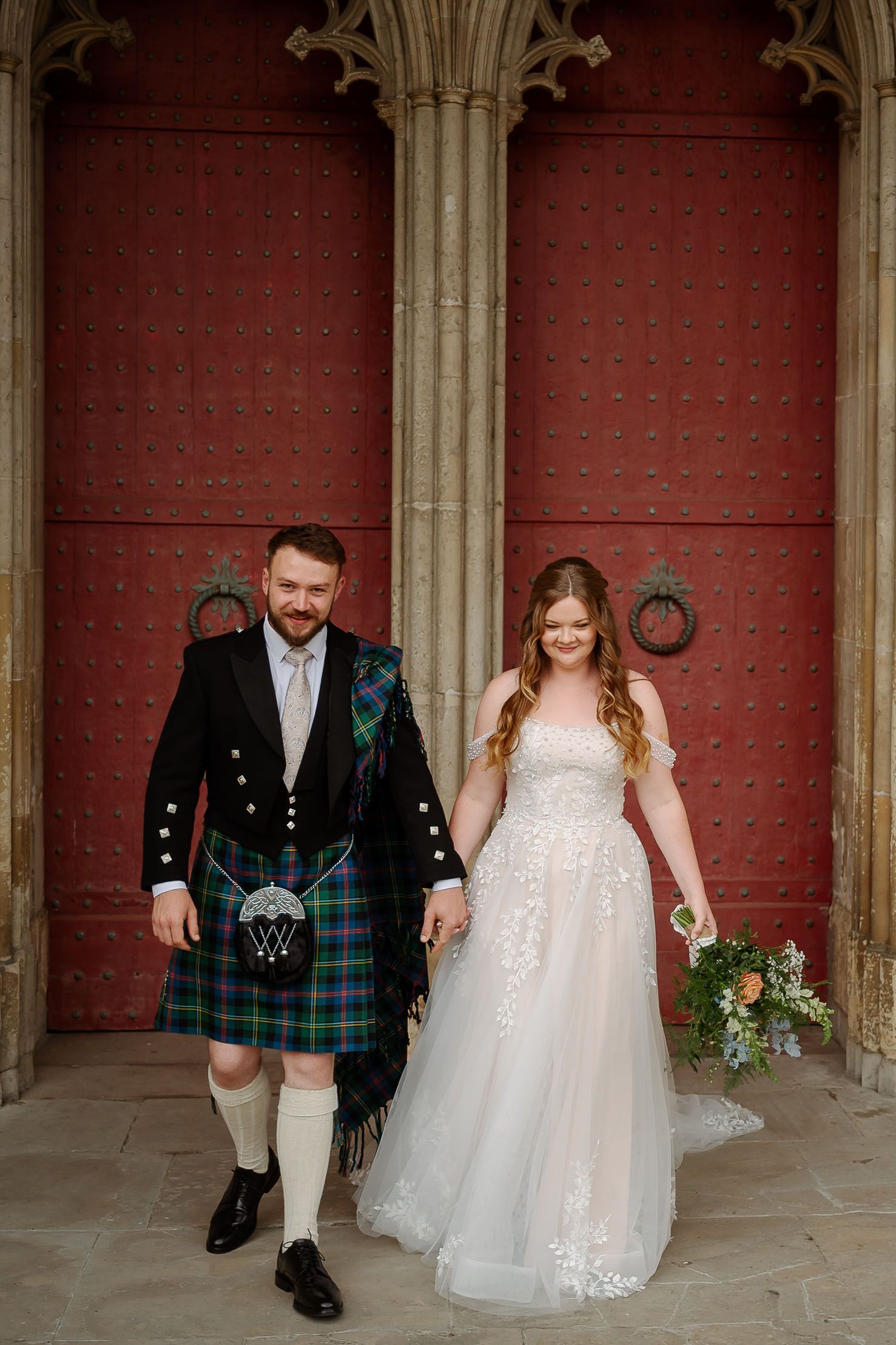 Bride and groom in Scottish kilt holding hands before red castle doors on their wedding day.