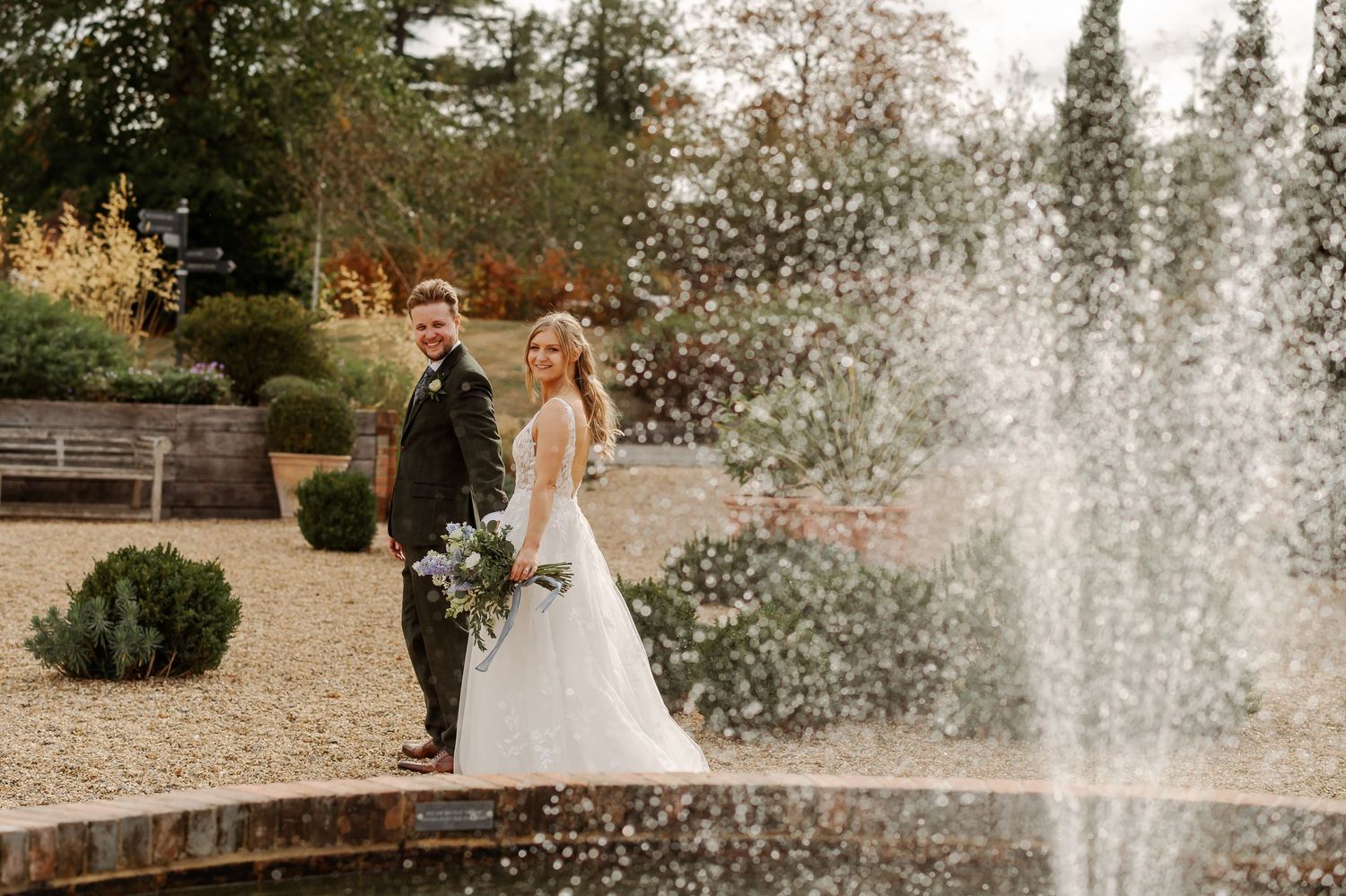 Bride and groom posing by a garden fountain on their wedding day, surrounded by lush greenery and autumn foliage.
