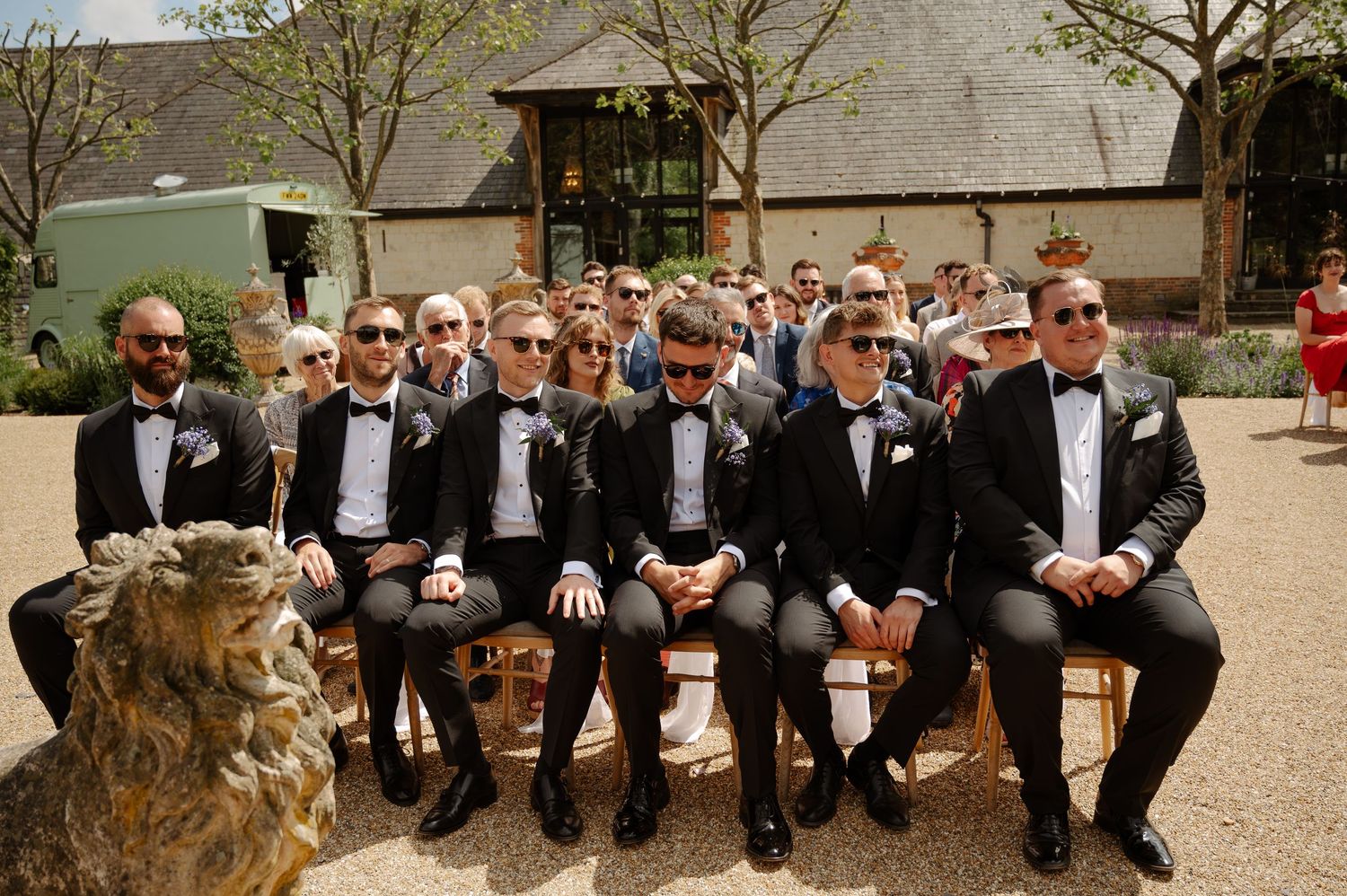 Groomsmen in black tuxedos sit casually on stone steps outdoors during a wedding photoshoot.