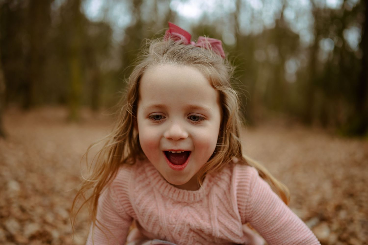 A joyful child in a pink sweater plays outdoors among fallen autumn leaves on a cloudy day.
