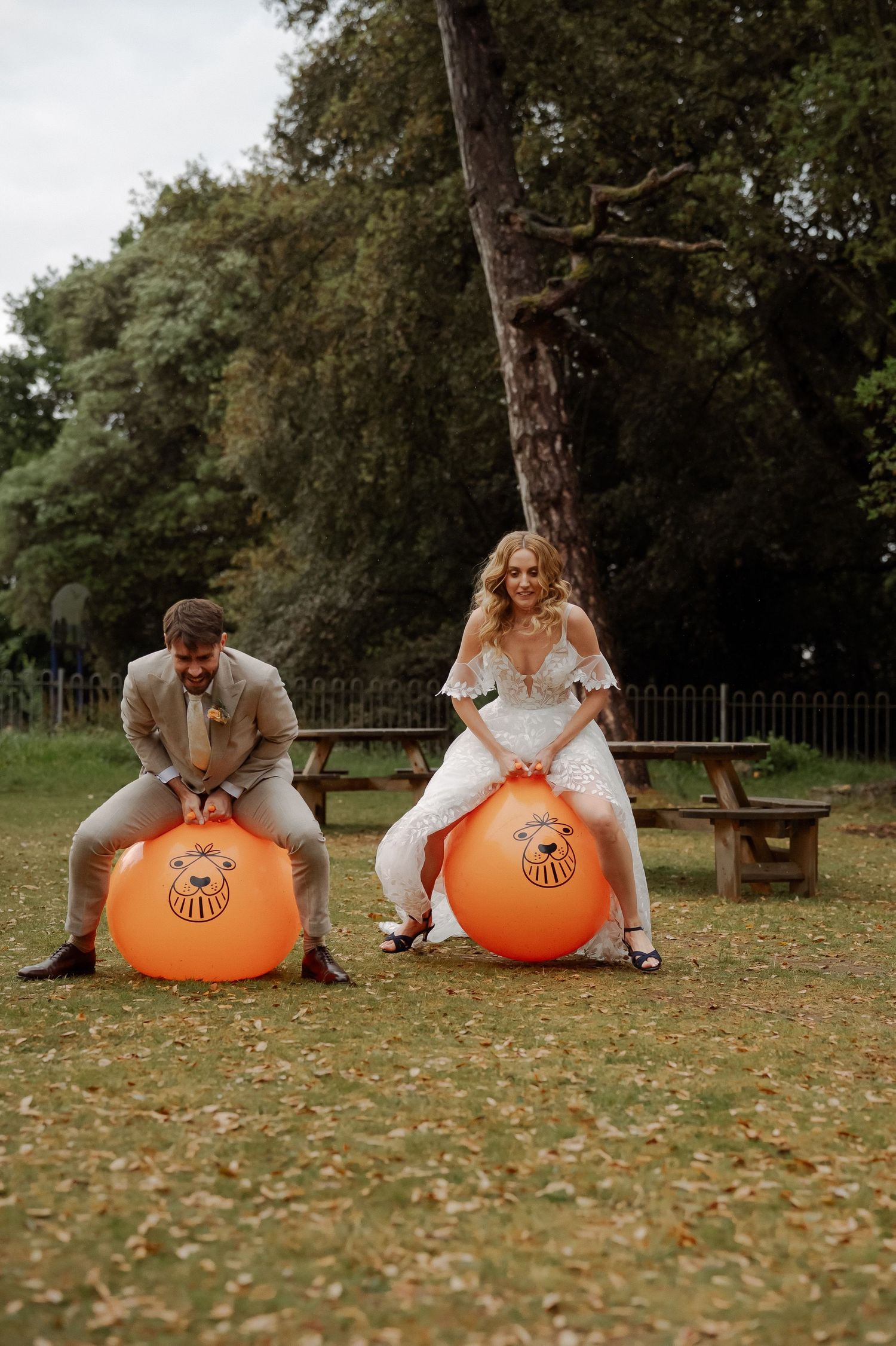 A couple sits back-to-back on orange bouncy balls in a park setting with fallen autumn leaves on the ground.
