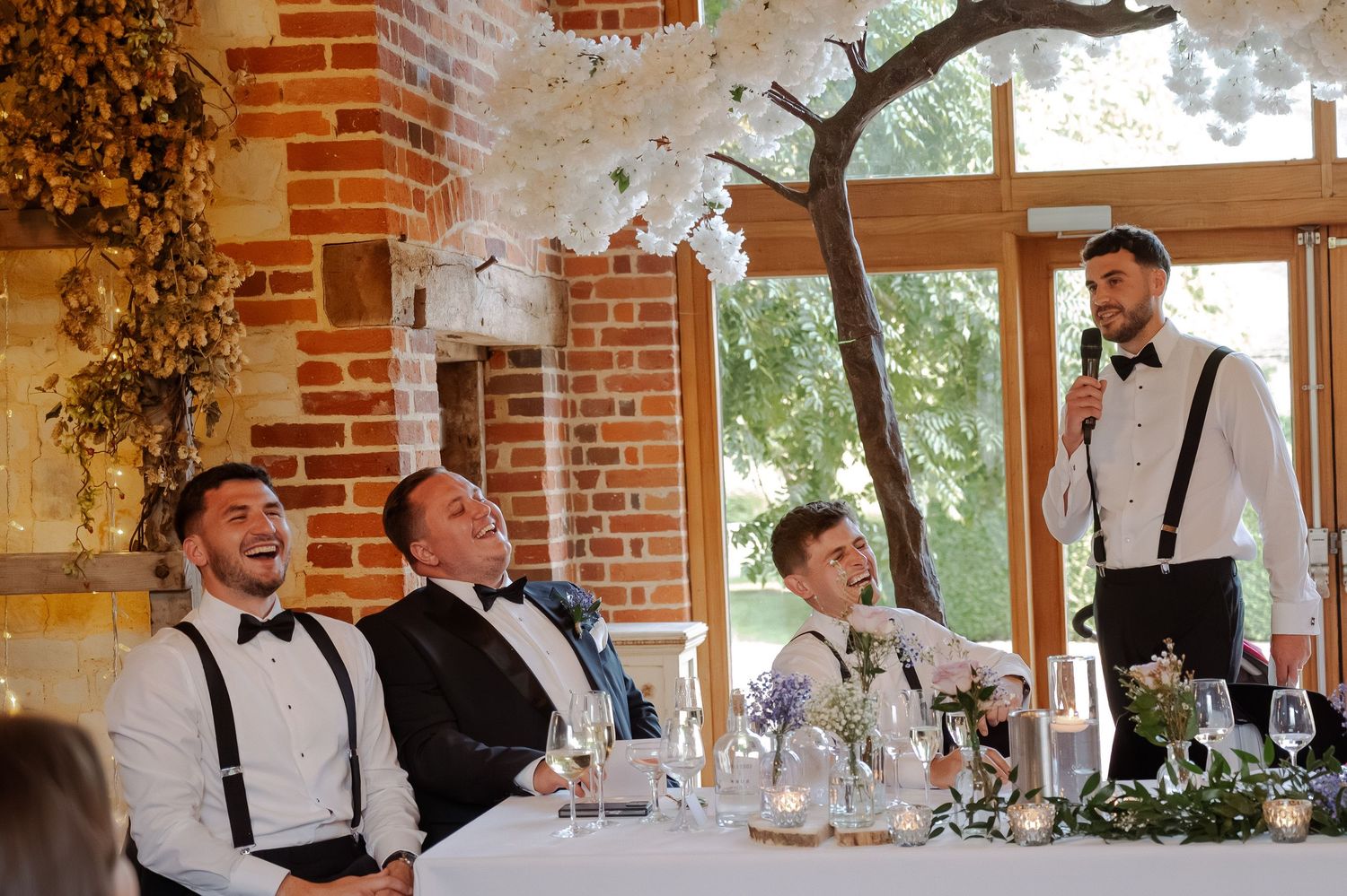 Wedding party members in suspenders and bow ties laugh and celebrate during reception at rustic brick venue.