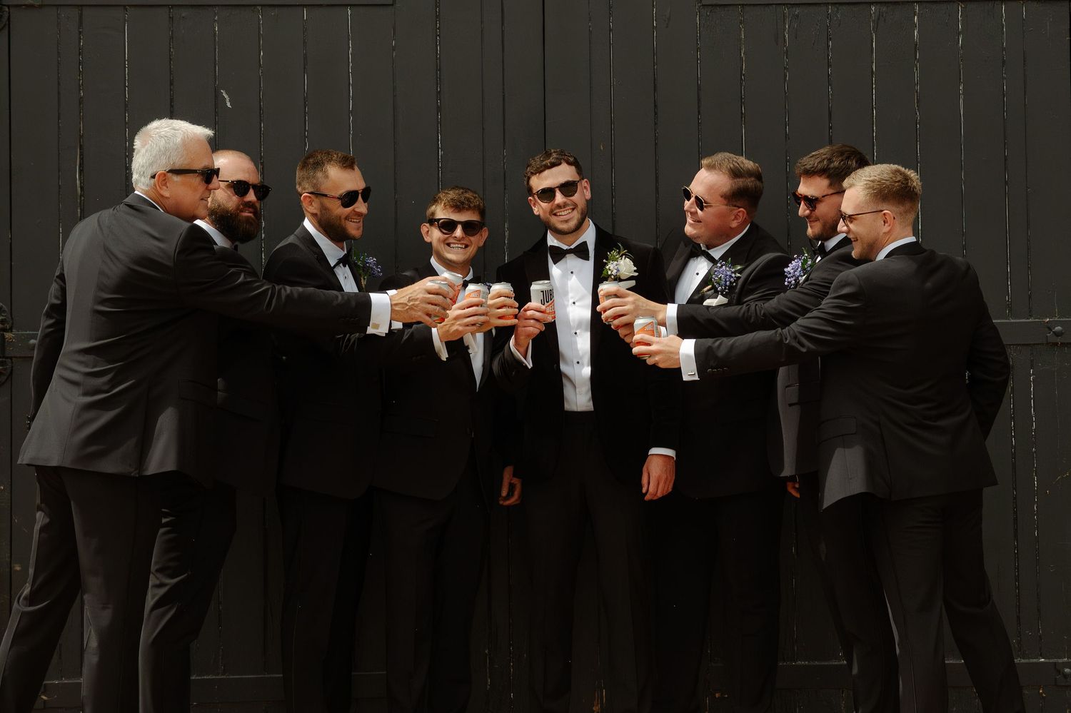 Groomsmen in black tuxedos raise glasses in a celebratory toast against dark wall at wedding celebration.