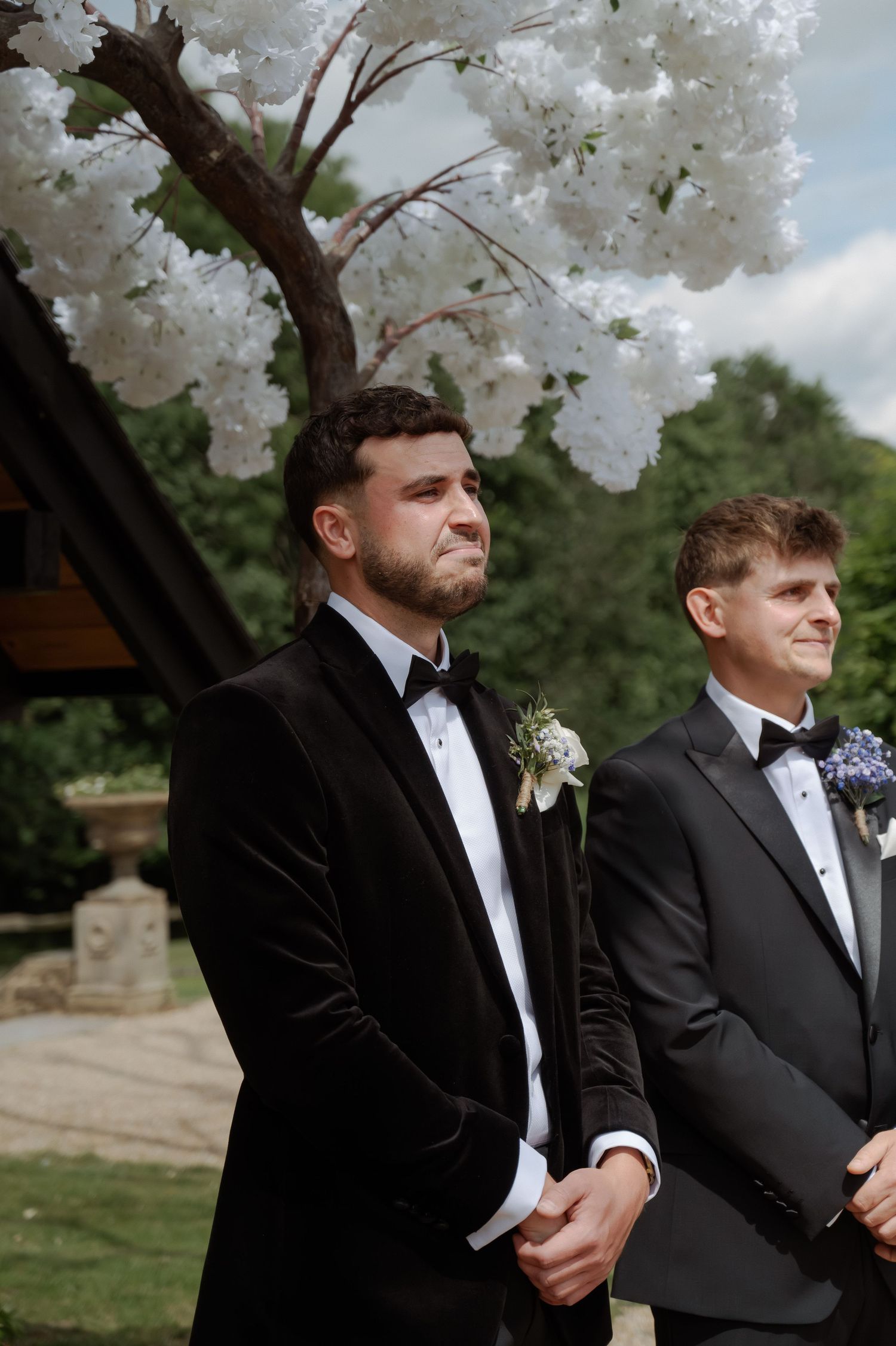 Two men in formal black tuxedos stand together outdoors under white flowering trees during a wedding ceremony.