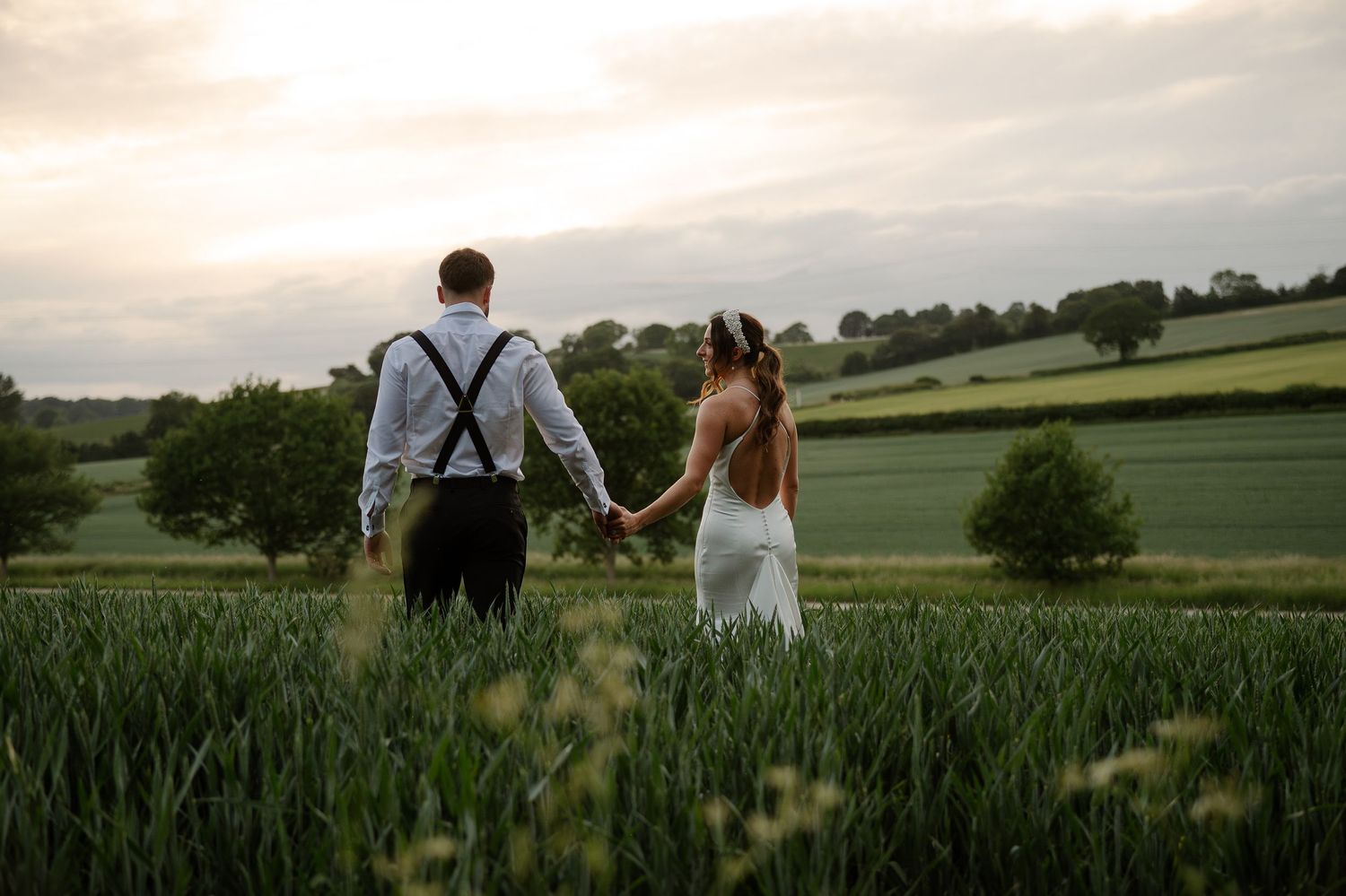 A couple holds hands while walking through a lush green countryside field at sunset.