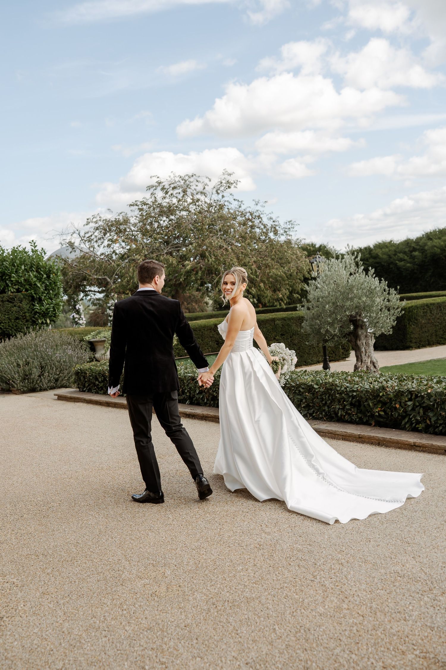 Bride in white gown and groom in black tuxedo walk hand-in-hand through elegant garden grounds on wedding day.