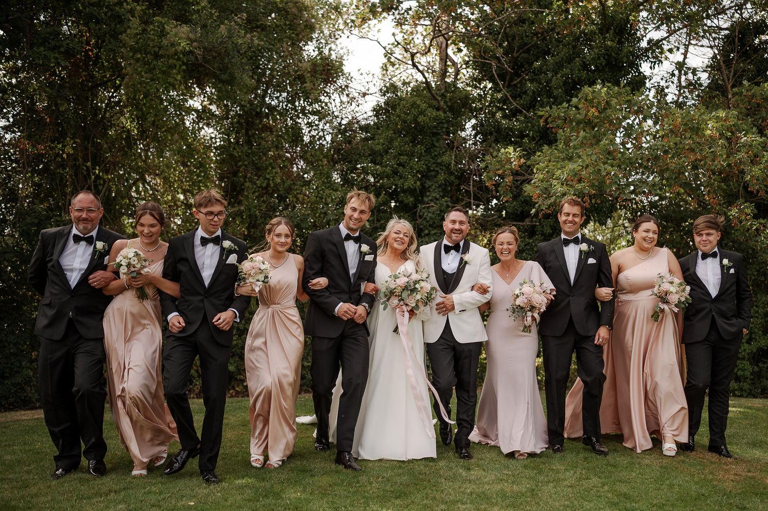 Wedding party in formal attire, bridesmaids in blush gowns and groomsmen in black tuxedos, posing outdoors with lush greenery.