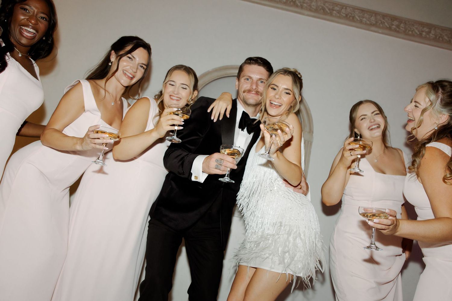 Bride in white feathered dress and groom in tuxedo celebrate with bridesmaids in blush gowns, holding champagne glasses.