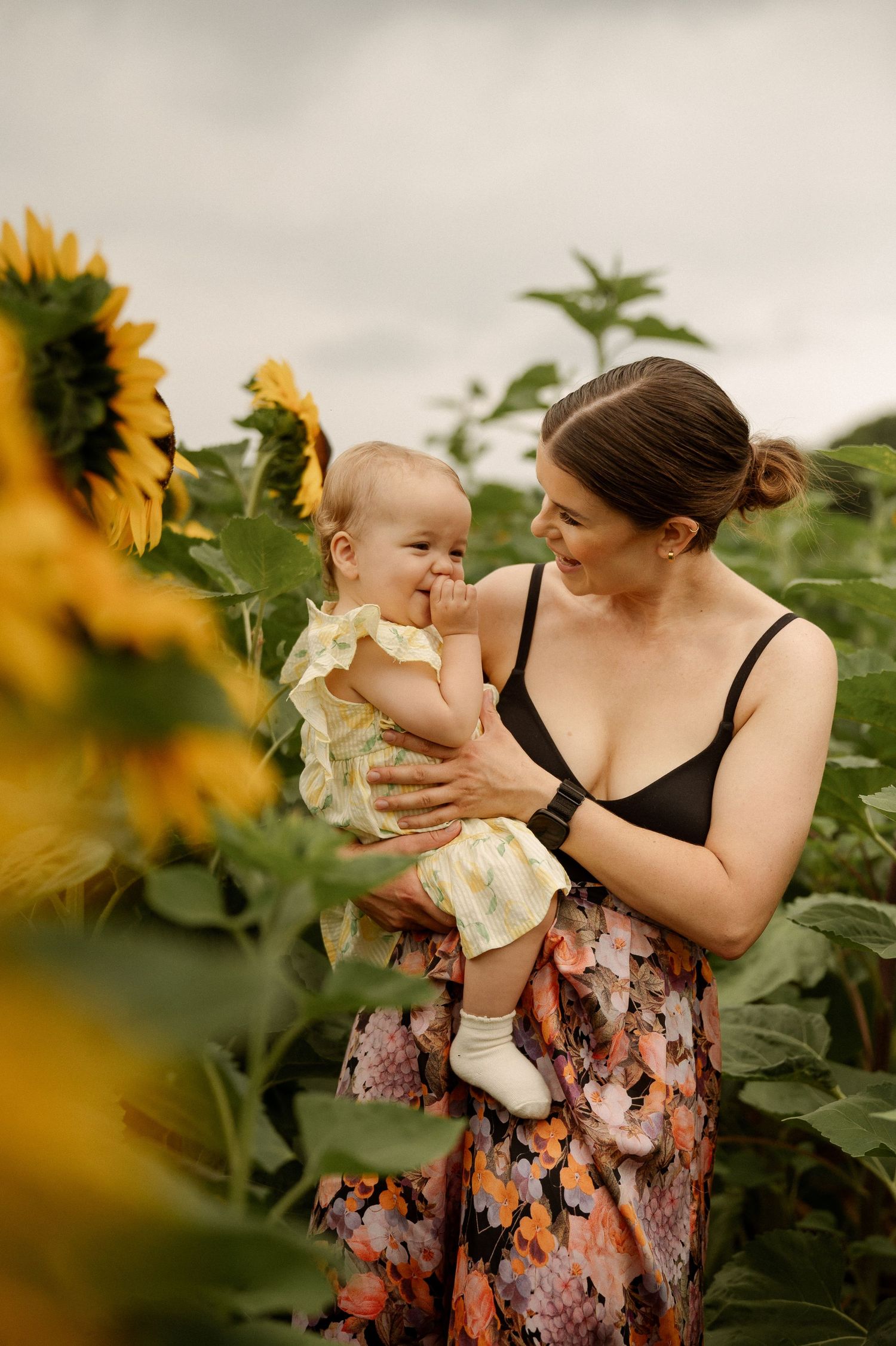 A mother holds her smiling baby in a vibrant sunflower field, wearing a floral skirt and black top on a cloudy day.