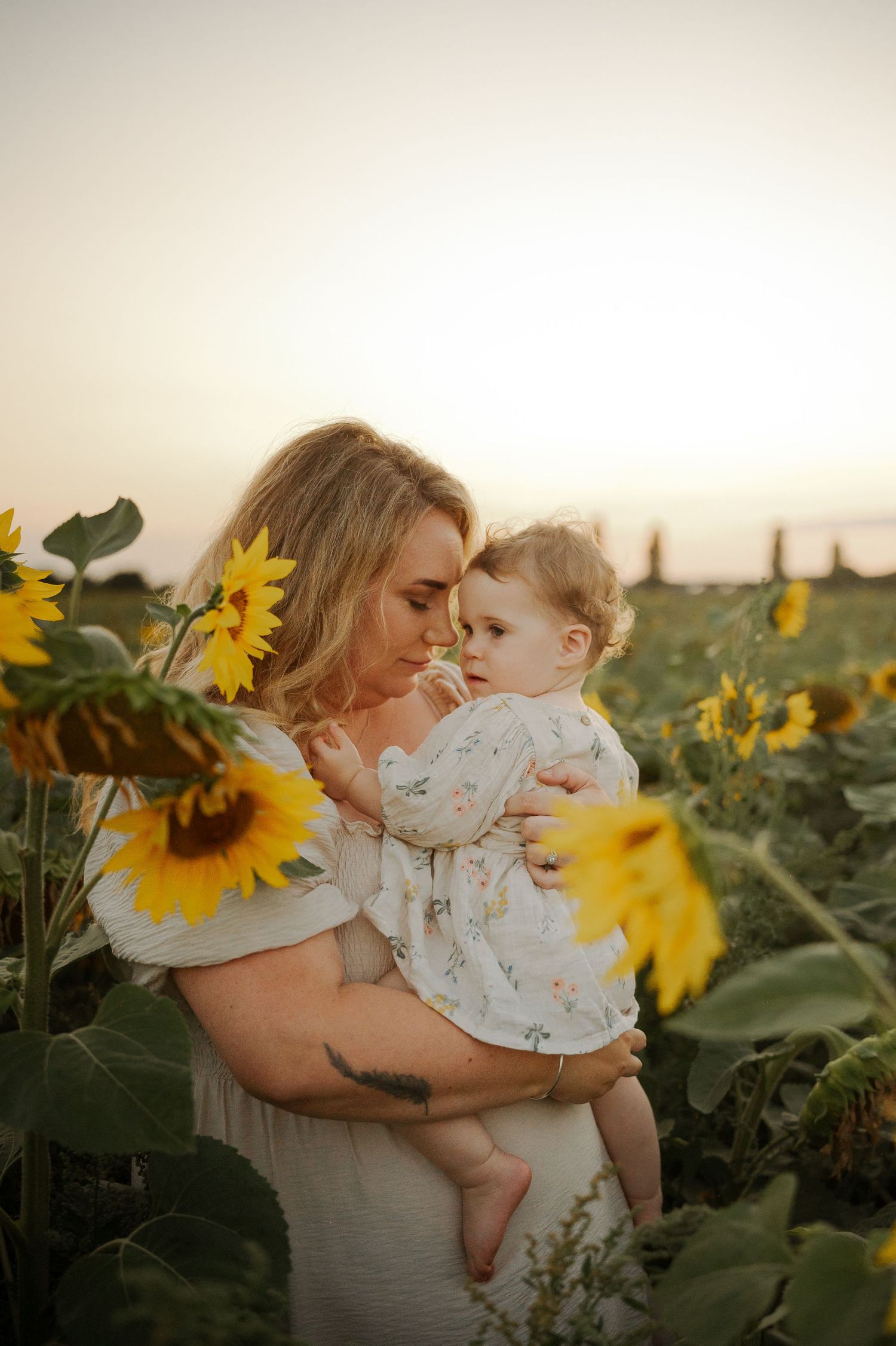 A mother holds her baby in a sunflower field at golden hour, both wearing white in a tender outdoor portrait.