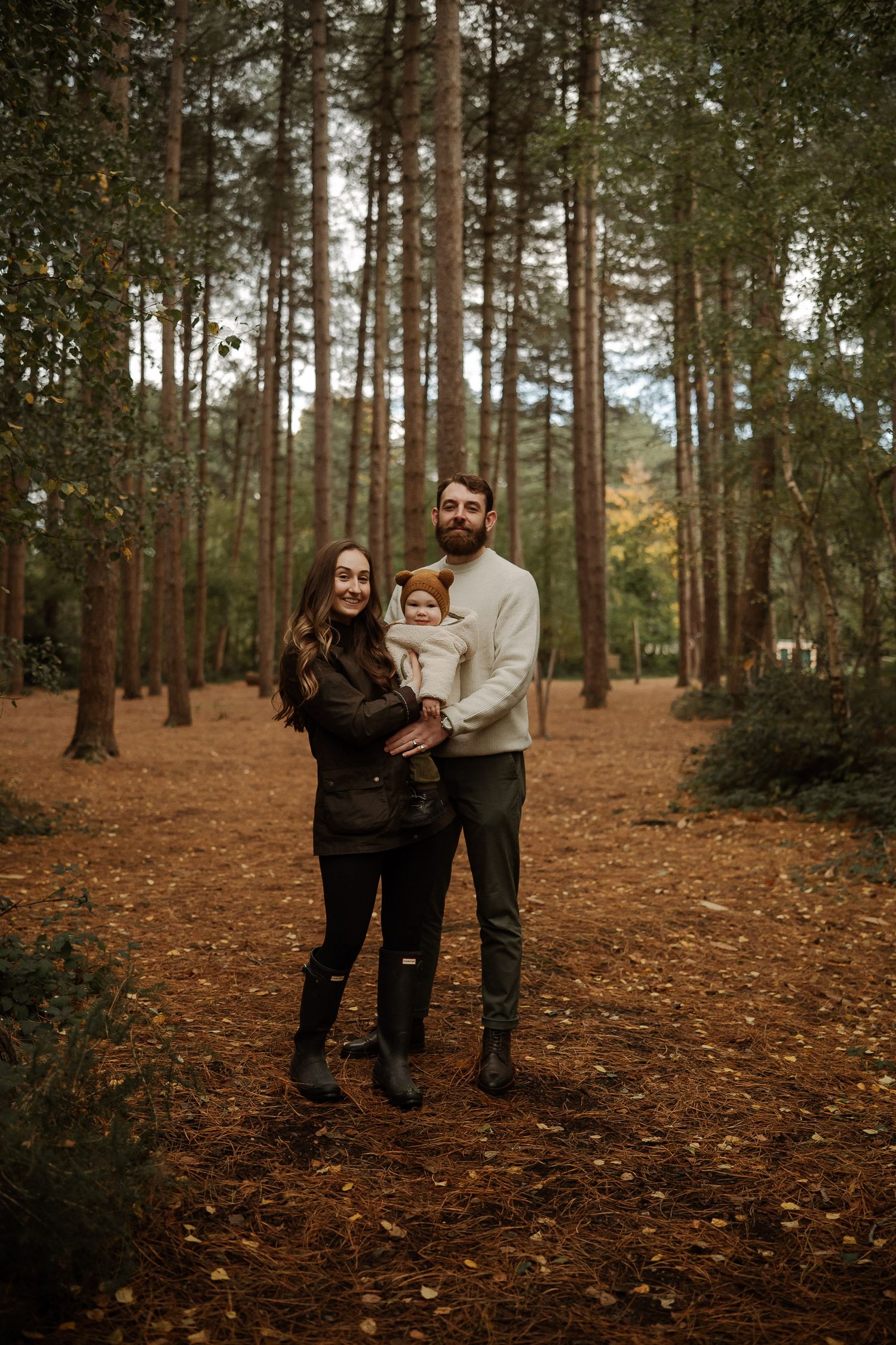 A couple walks together through a serene autumn forest with pine trees and fallen orange leaves covering the ground.