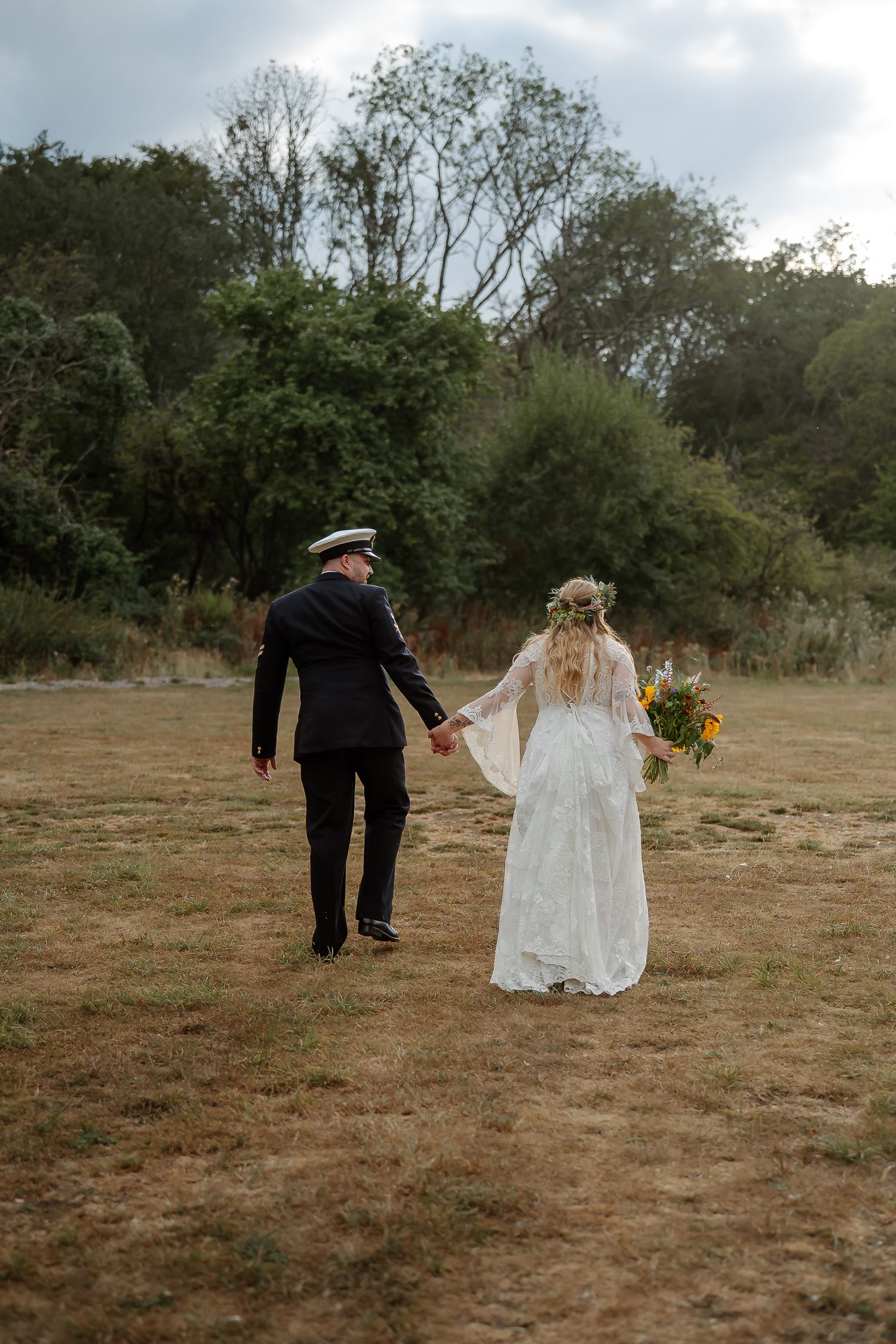Bride and groom walking hand in hand through a rustic field, bride holding sunflowers in a lace gown, groom in military dress uniform.