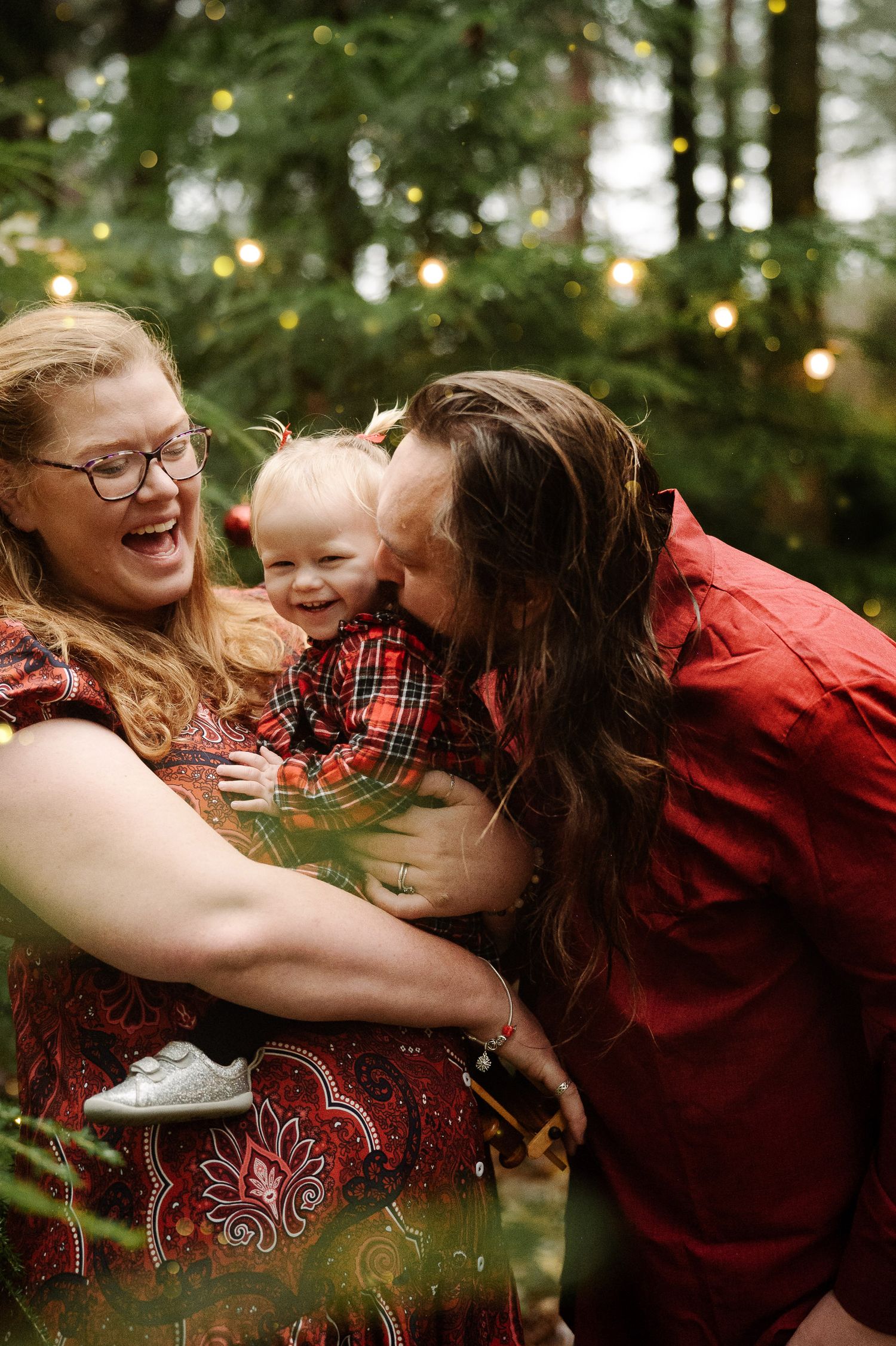 Joyful family with parents kissing laughing toddler outdoors among festive bokeh lights in red holiday outfits.