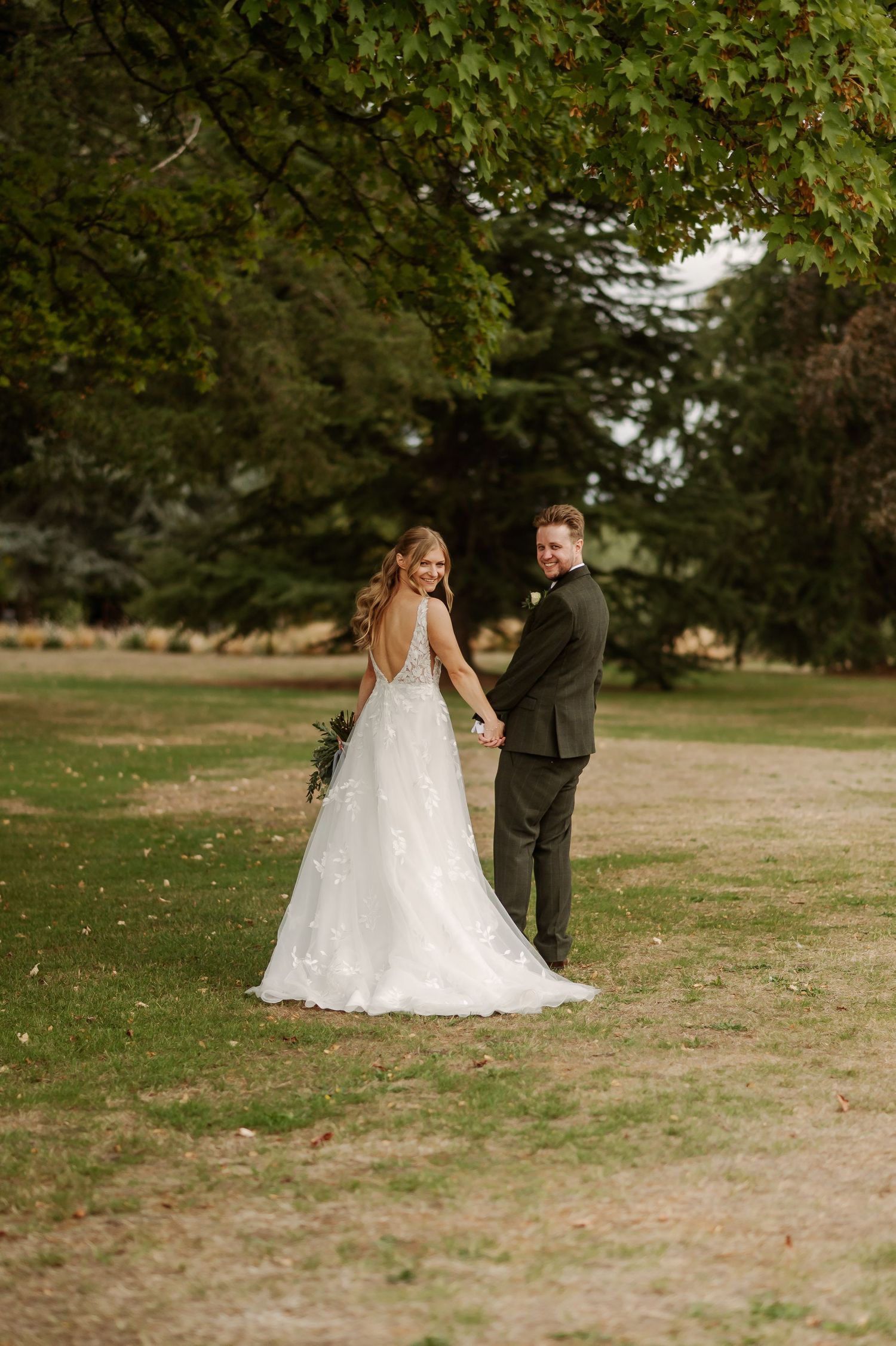 Bride and groom holding hands walking through lush green park, bride in elegant backless white gown with floral train.
