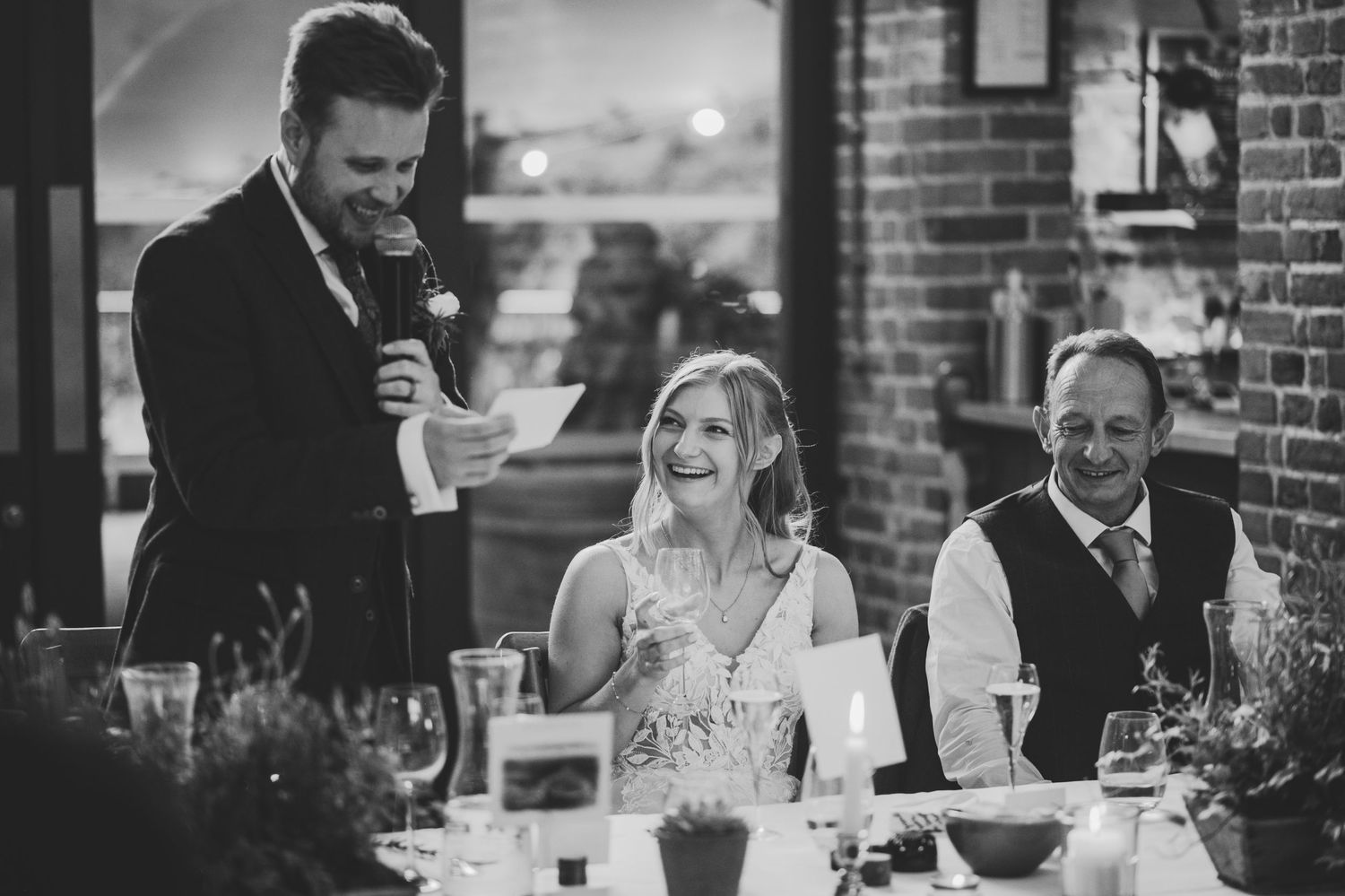 Black and white wedding reception photo of groom giving speech while bride and guest laugh joyfully at elegant dinner table.