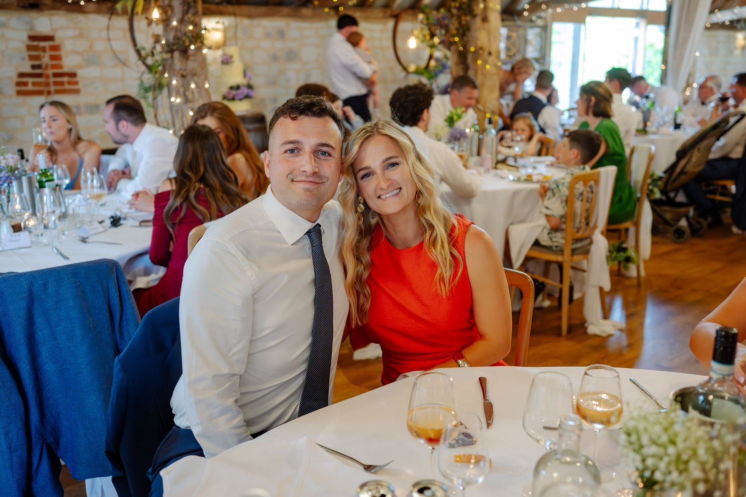 Wedding guests enjoying a meal and conversation at decorated tables in a rustic indoor reception space.