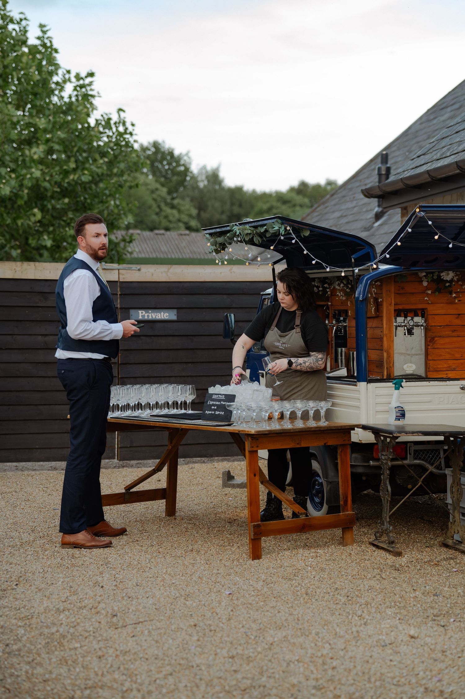 Person setting up a rustic outdoor drink station with wooden tables and glassware.