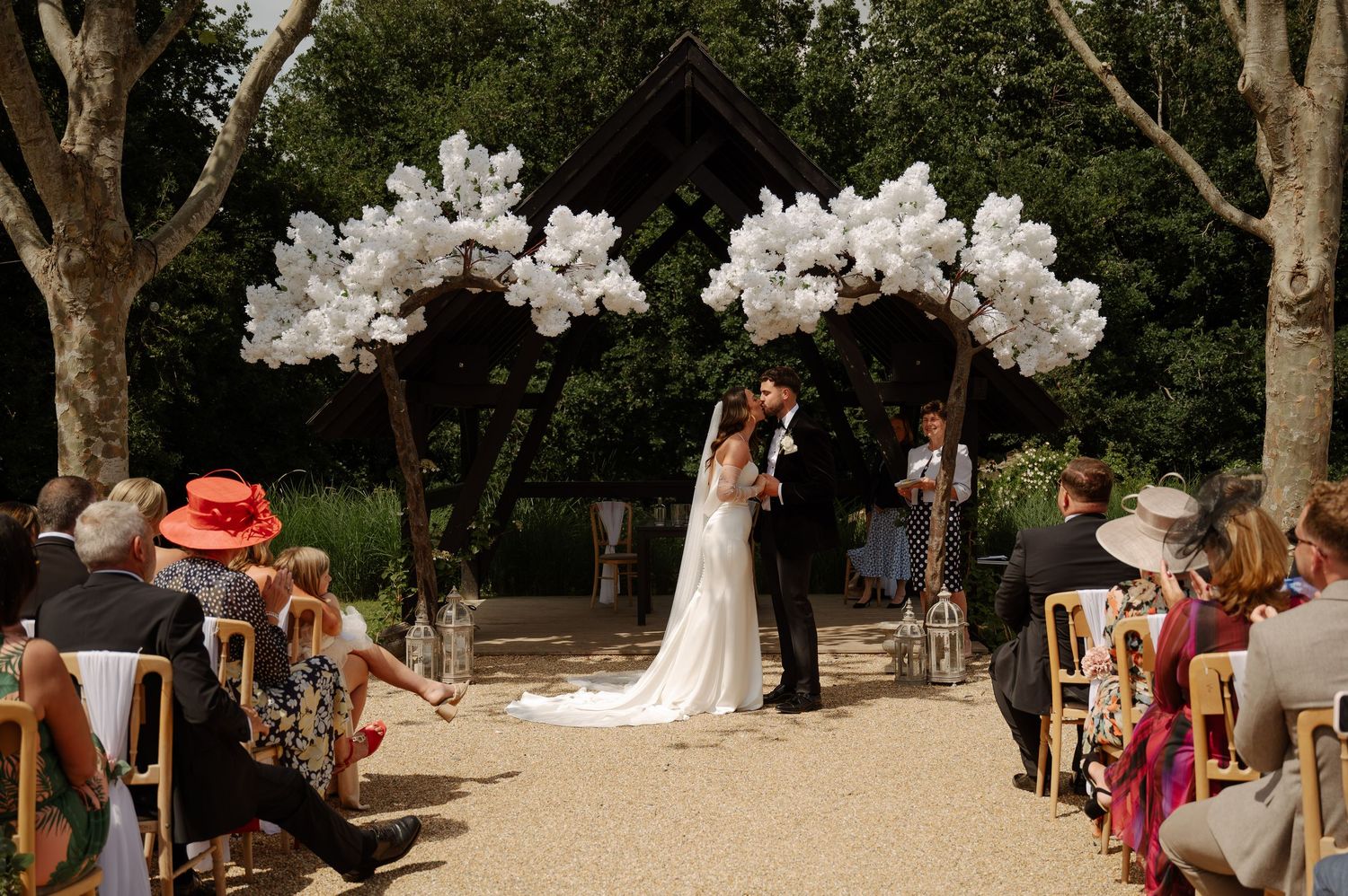Wedding ceremony setup outdoors with white floral arrangements and guests gathered under tall trees.