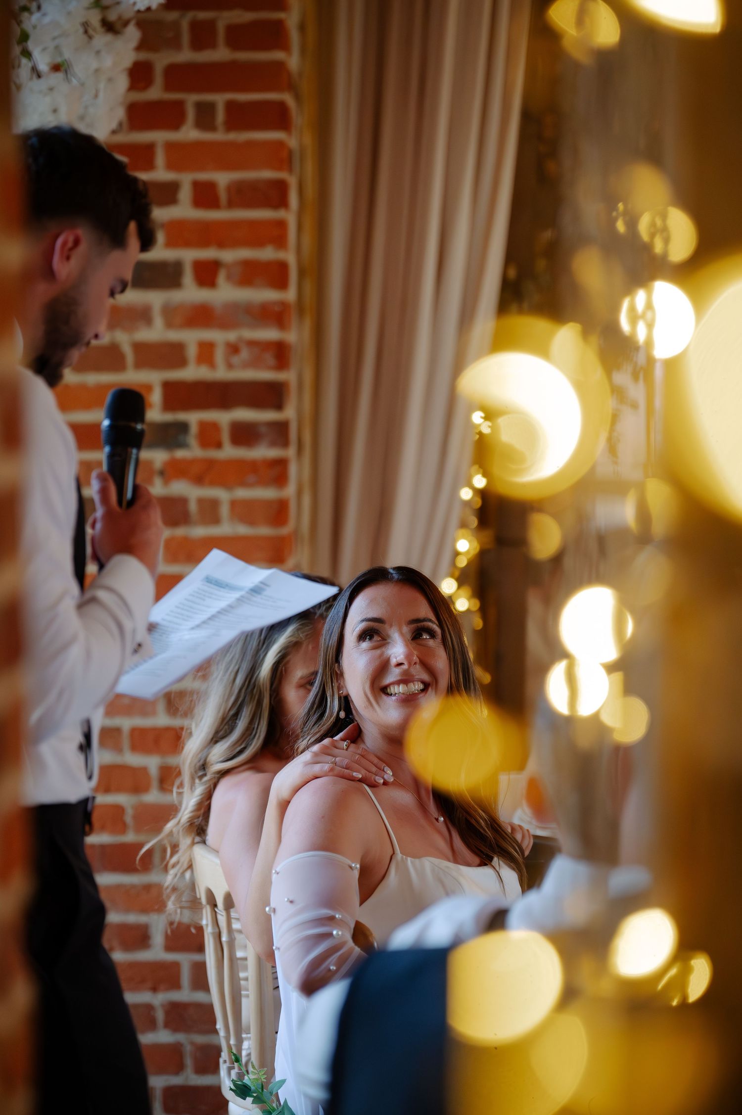 Wedding guests dance and celebrate at an indoor reception with exposed brick walls and warm string lights creating ambiance.