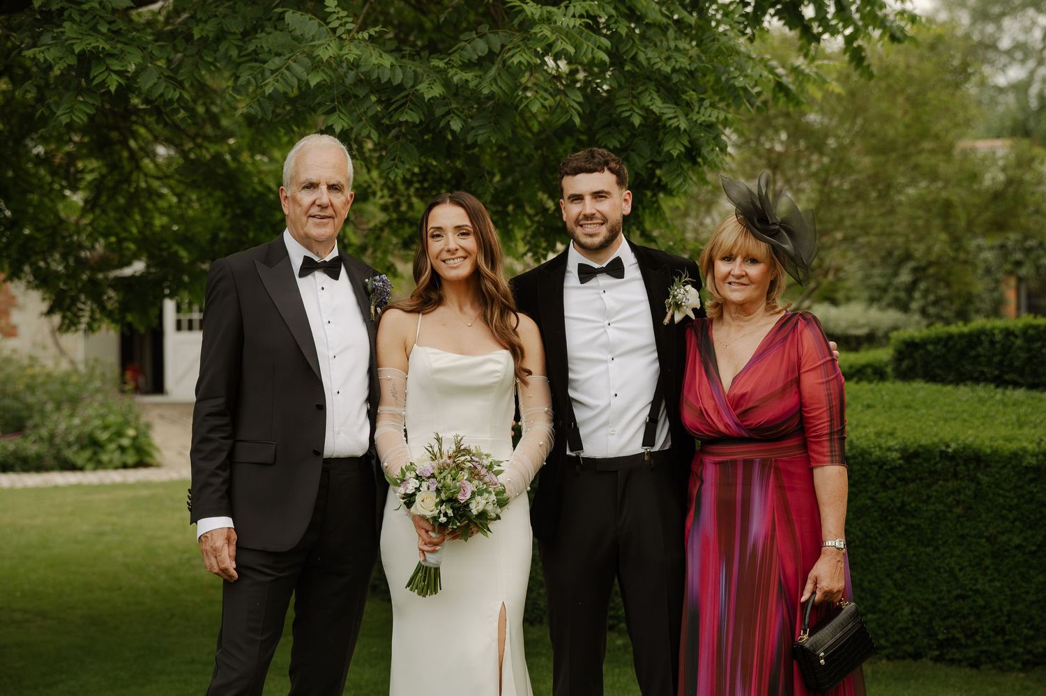 A formal group poses together outdoors on a lawn with green hedges and trees in the background.