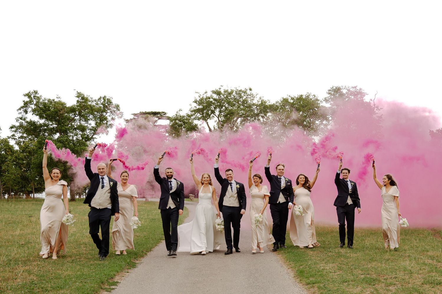 Wedding party celebrates on outdoor path with vibrant pink smoke bombs creating a festive atmosphere.
