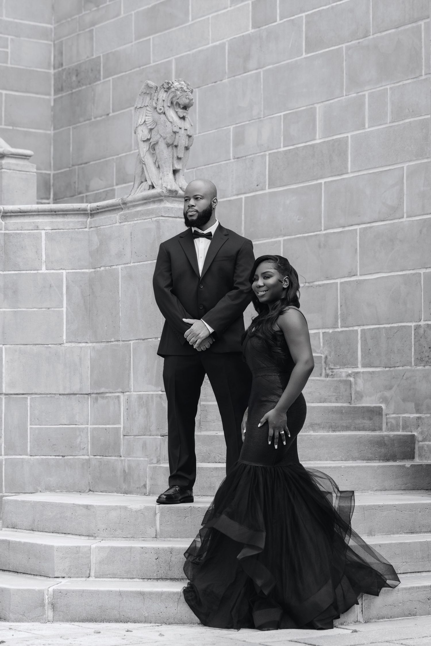 Black and white photo of couple in black formal attire posing on stone steps near angel sculpture.
