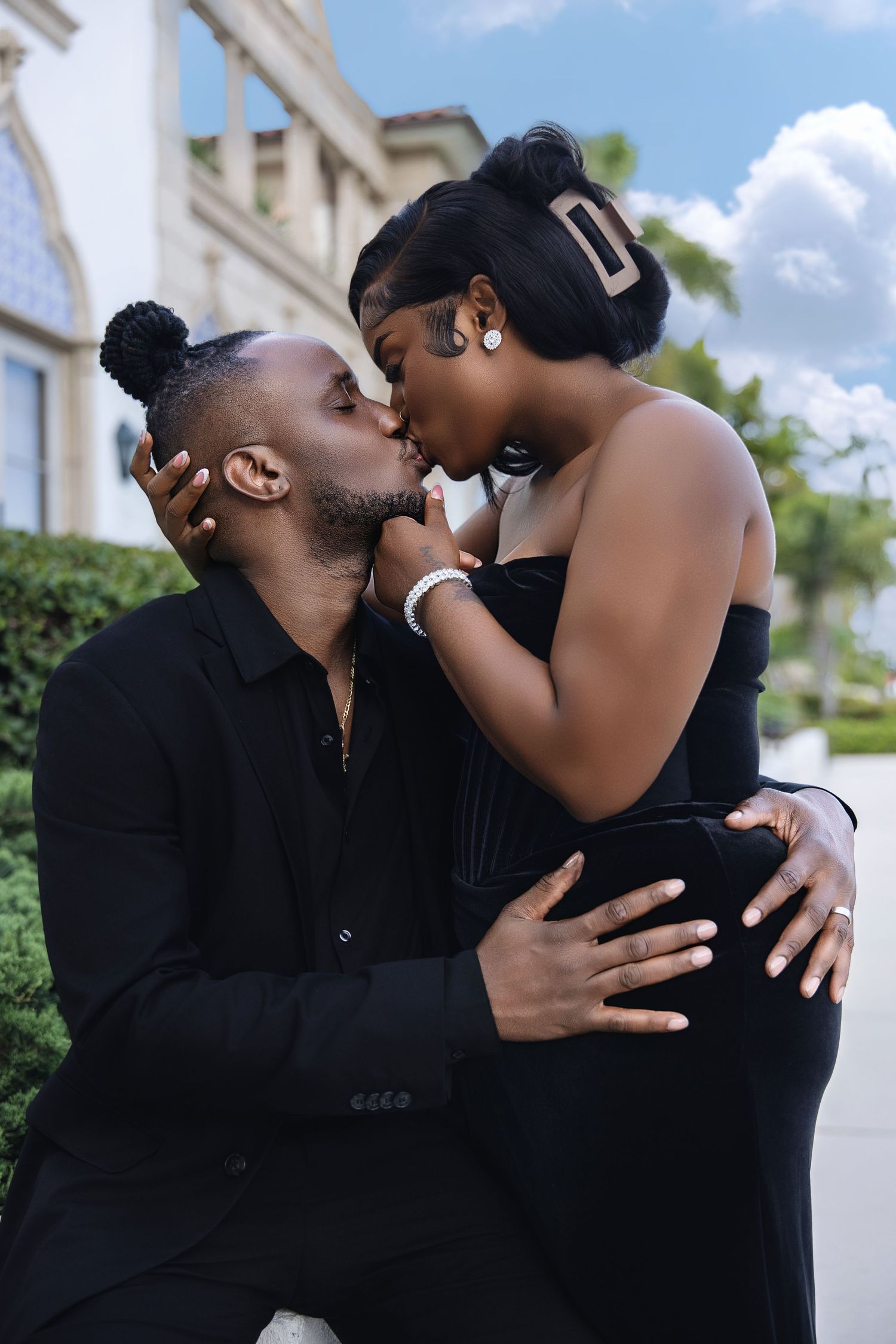 A Black couple shares a romantic kiss outdoors, both dressed elegantly in all-black outfits.