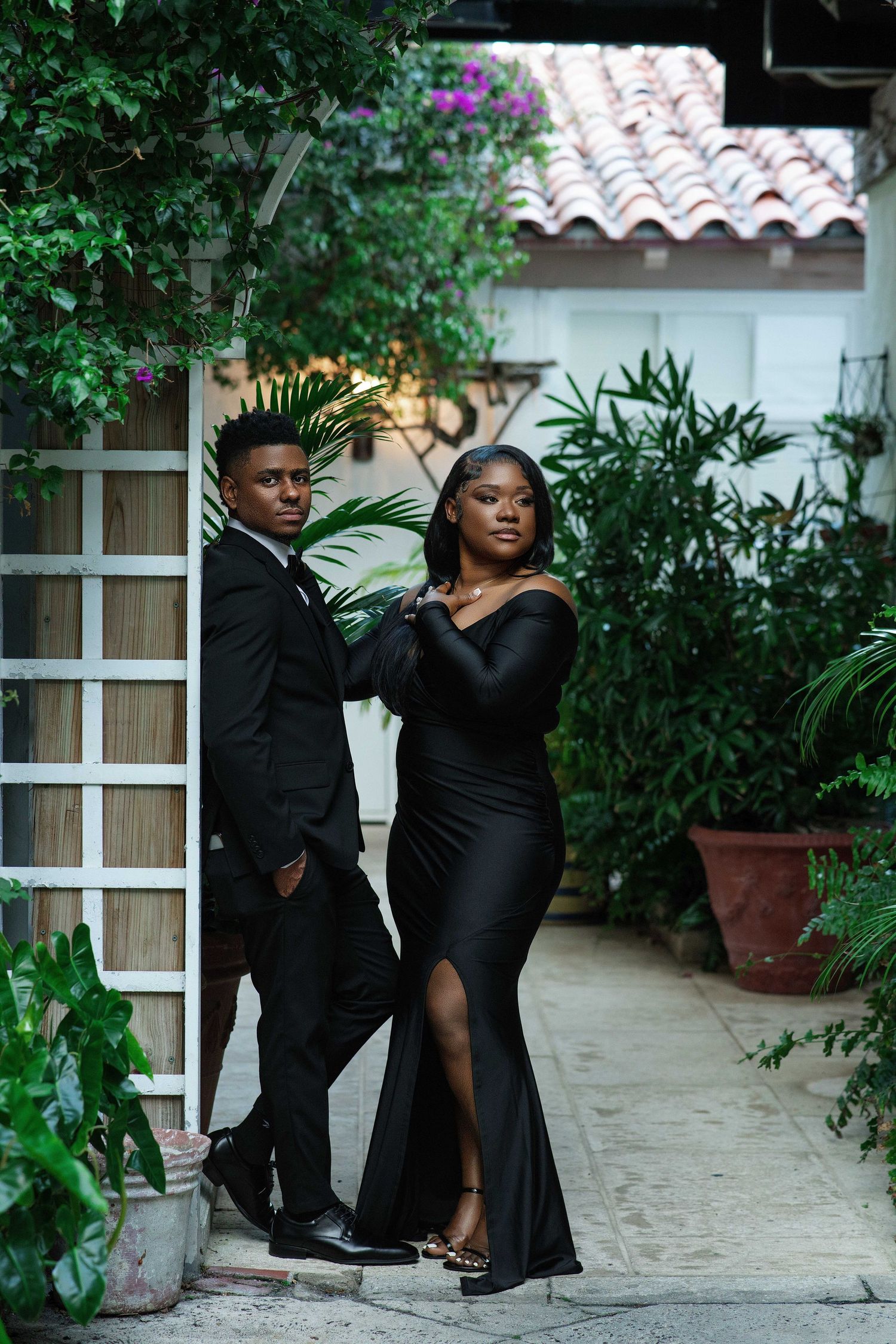 An elegant couple in matching black formal outfits pose together in a lush tropical garden setting.