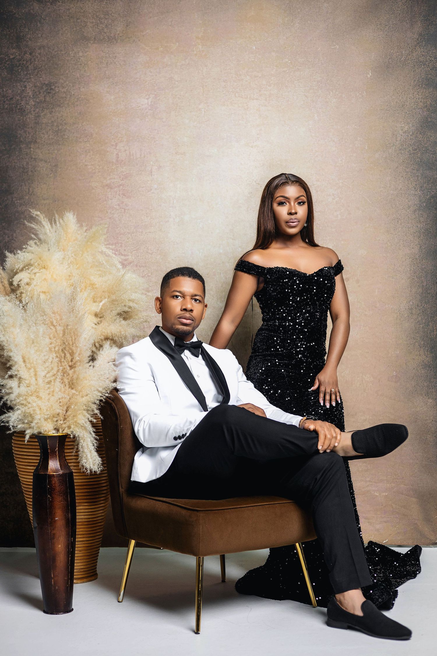 Sophisticated couple posing in studio, man in white tuxedo seated, woman in black sequin gown.
