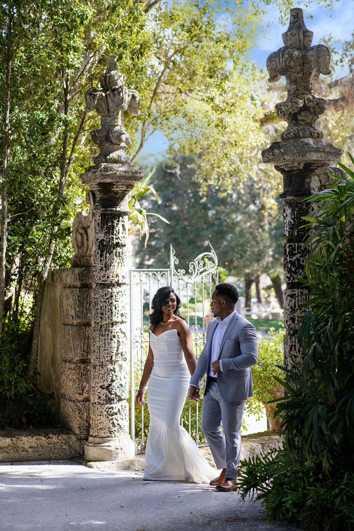 Bride in white mermaid gown and groom in gray suit walking through ornate stone gate.