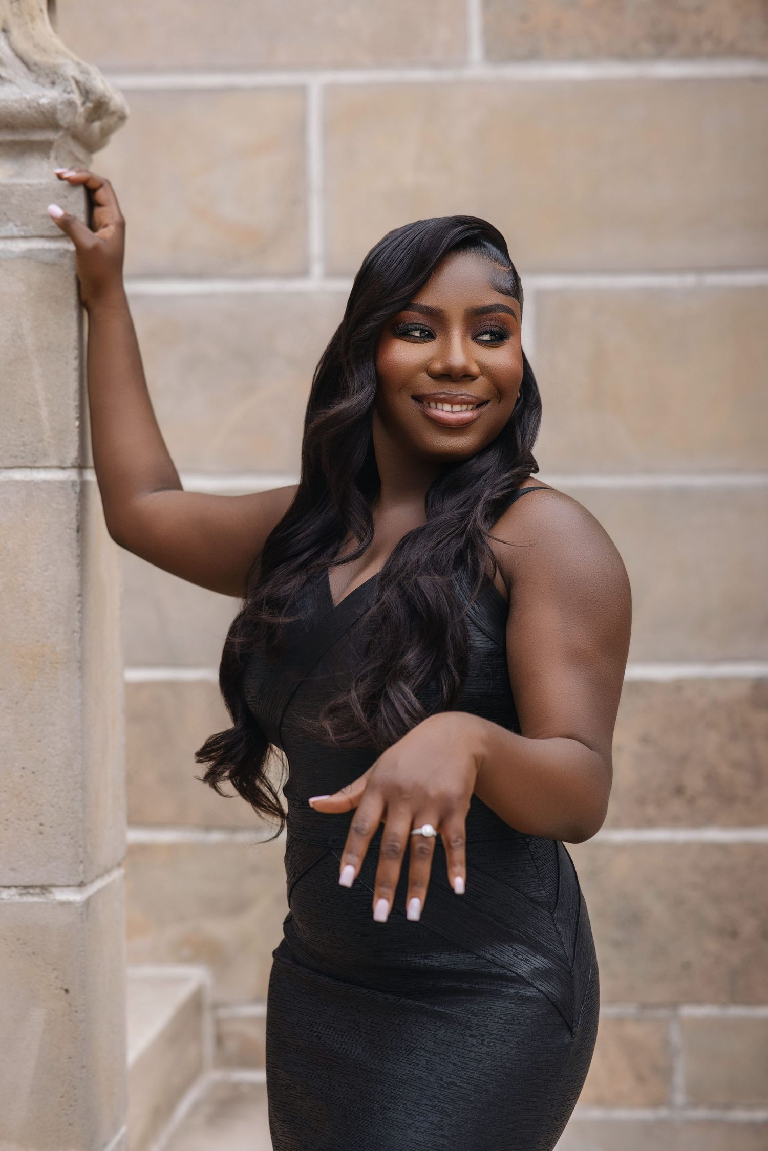 Beautiful woman in black dress smiling and displaying engagement ring while leaning against stone wall.