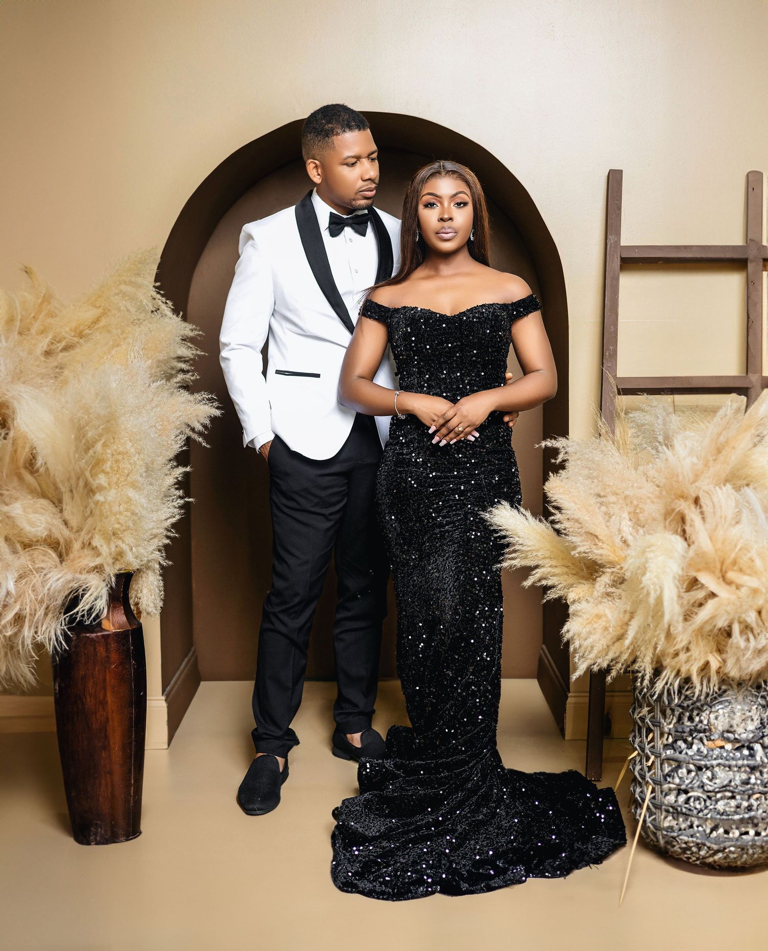 Couple in elegant black and white formal attire posing in studio with pampas grass backdrop.