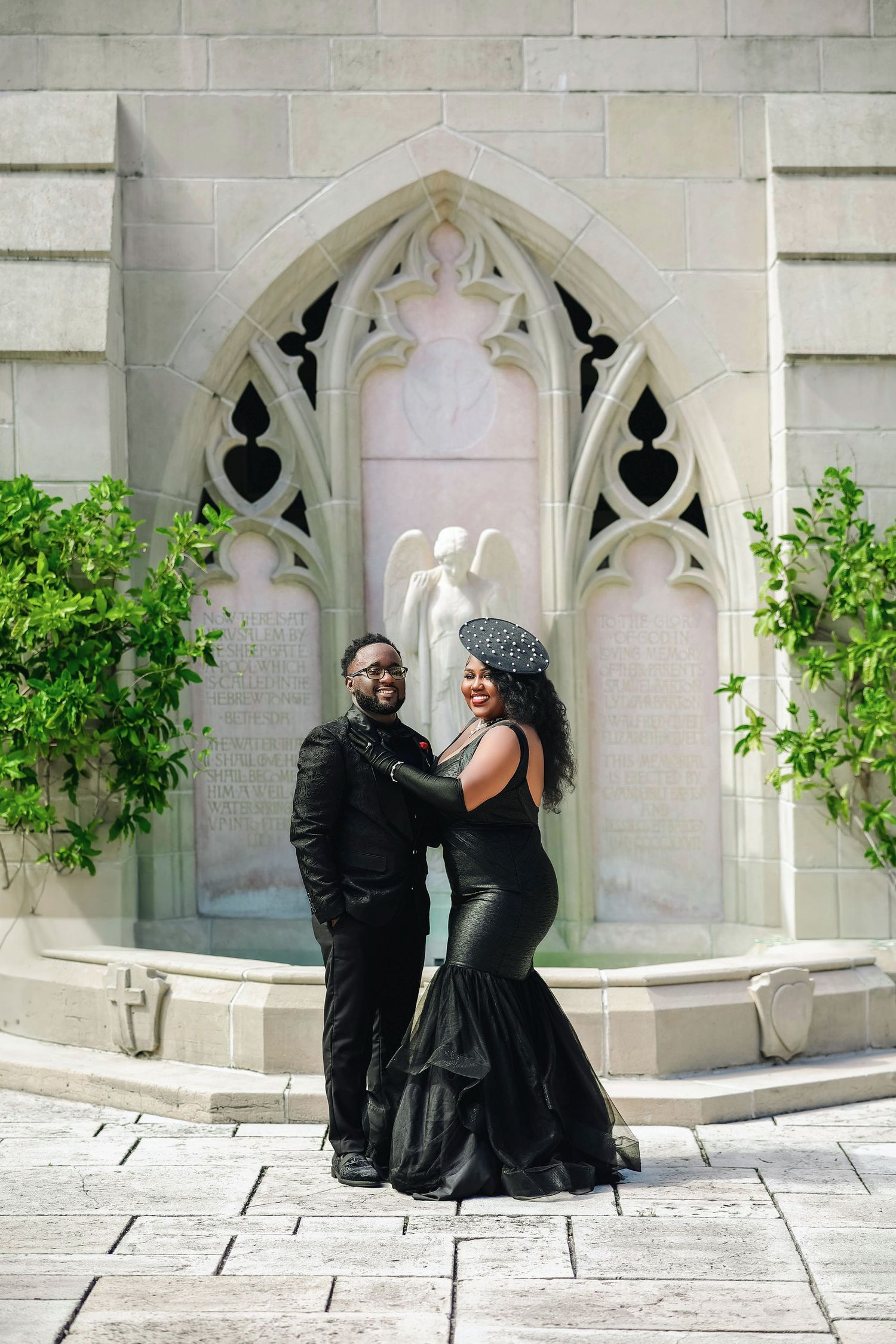 A couple in elegant black formal wear pose together before a Gothic stone archway with angel sculpture.