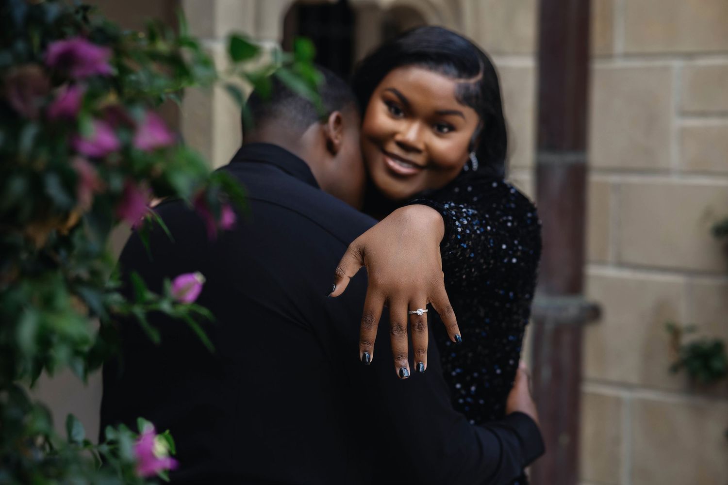 Smiling woman showing engagement ring while embracing her partner near flowering vines and stone wall.