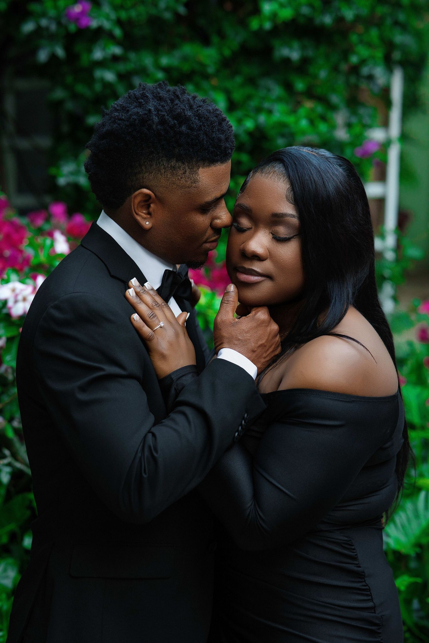 A couple in elegant black formal attire share a tender, close embrace surrounded by lush green foliage.