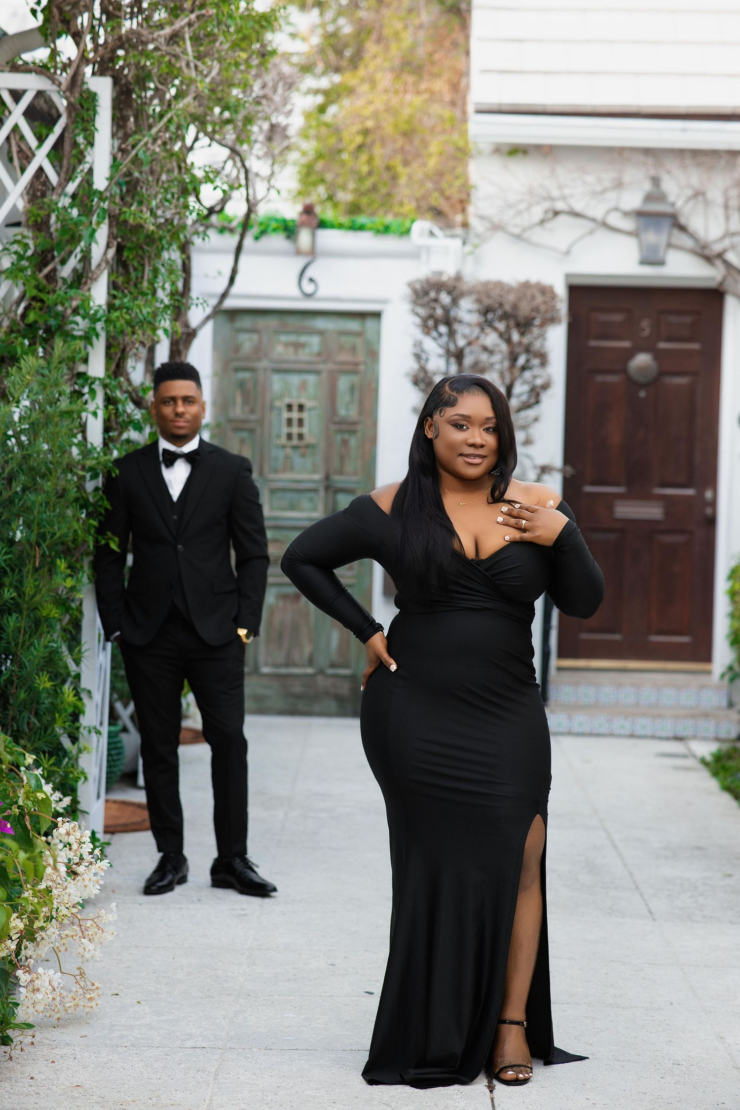 Woman in black off-shoulder gown posing with man in tuxedo in garden courtyard.