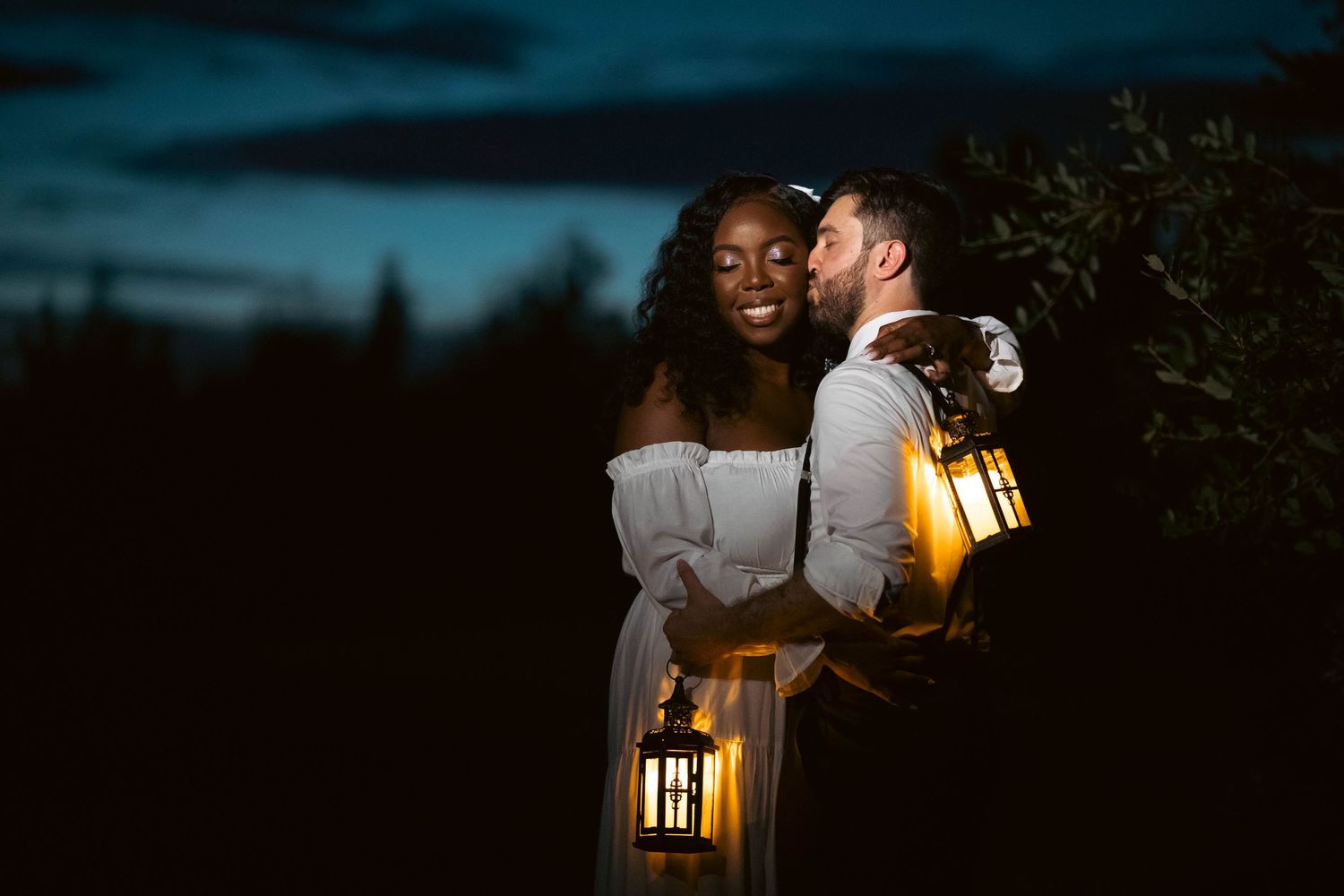 Couple holding glowing lanterns at dusk, sharing a romantic kiss outdoors at their wedding.