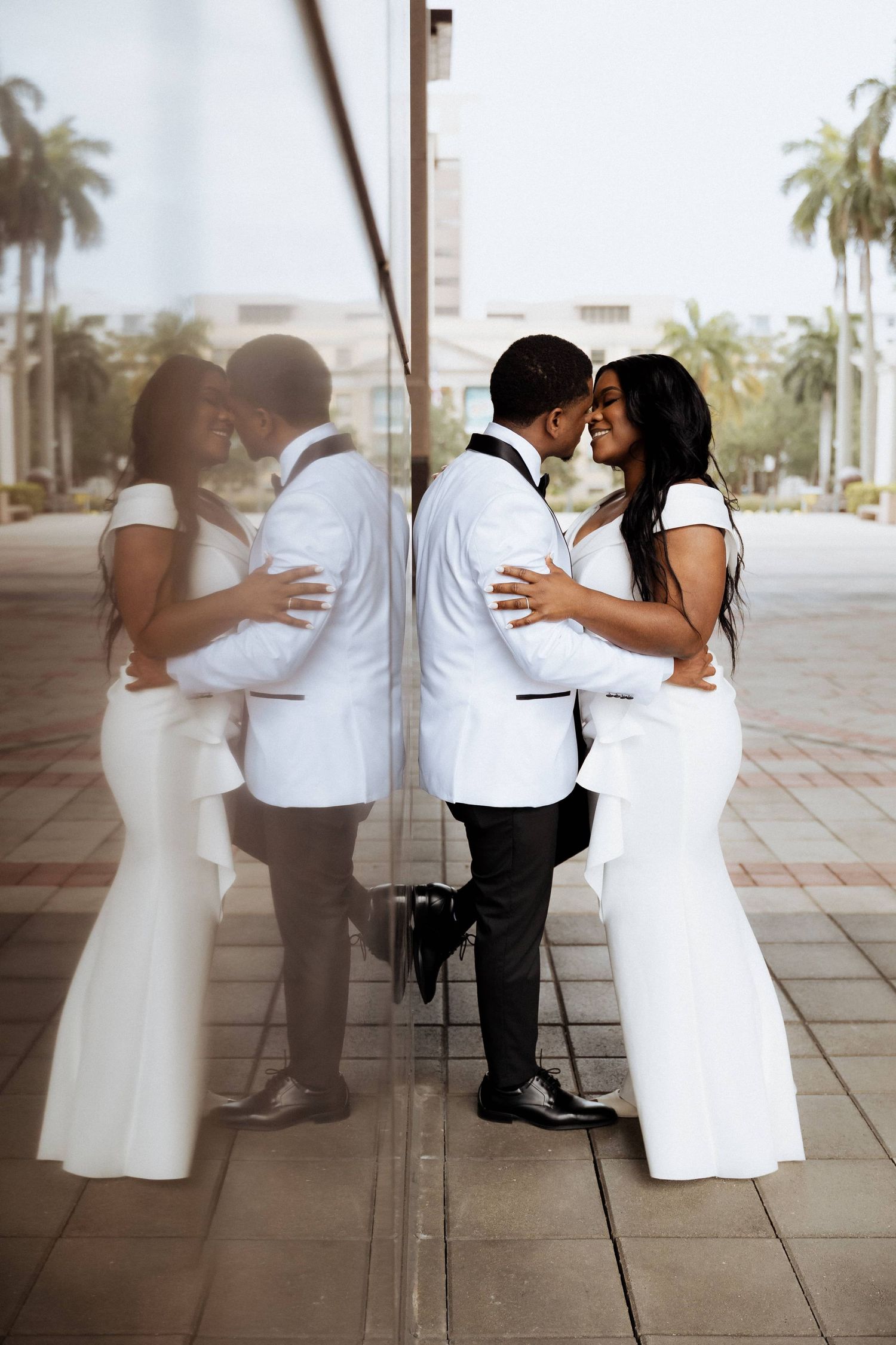 Elegant couple in wedding attire sharing a romantic moment, reflected in a mirrored wall outdoors.