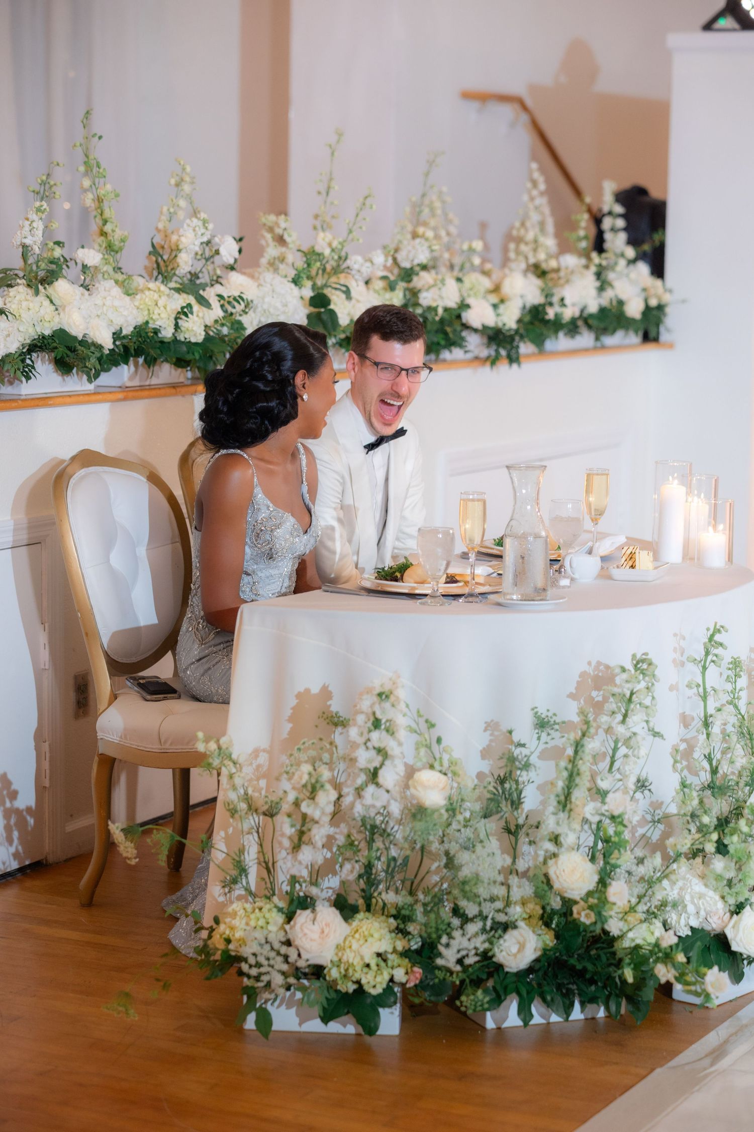 Bride and groom laughing at floral-adorned sweetheart table with white roses and candles at elegant wedding reception.