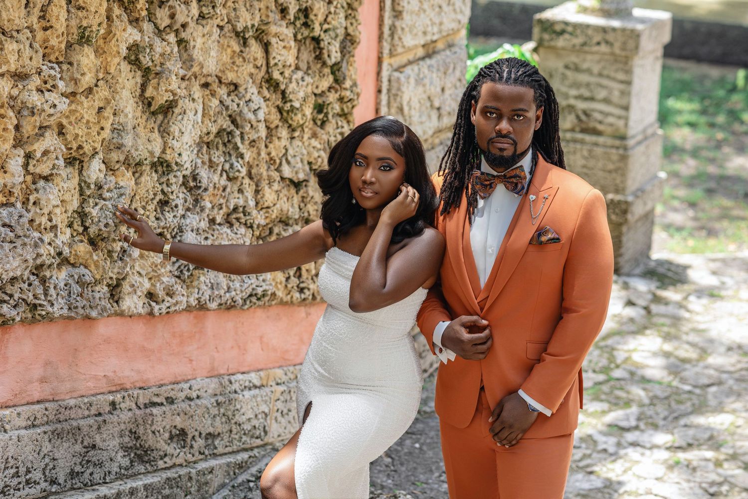 Couple in white dress and orange suit posing against a textured stone wall.
