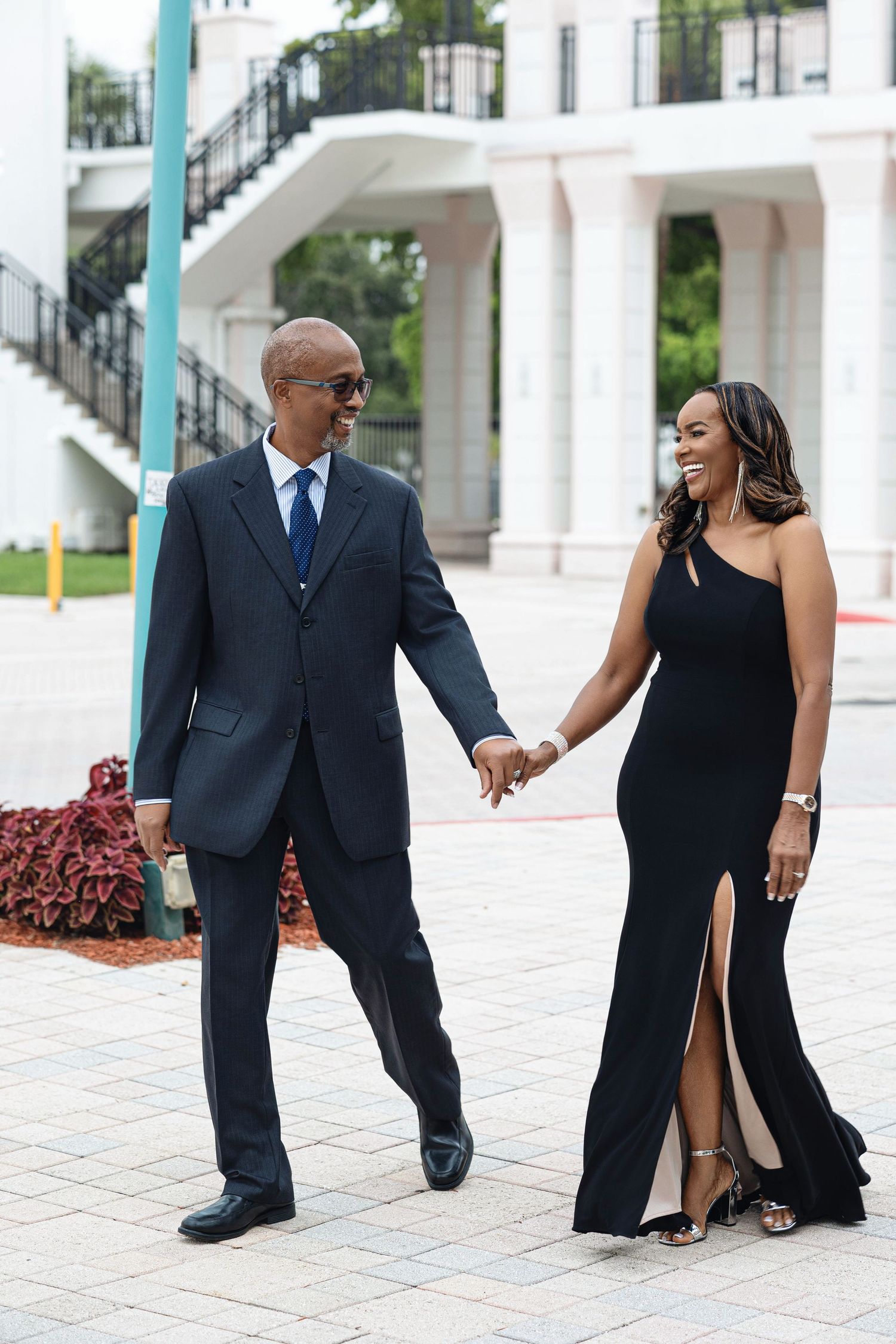 Happy engaged couple walking hand in hand outdoors, woman in elegant black gown, man in navy blue suit.