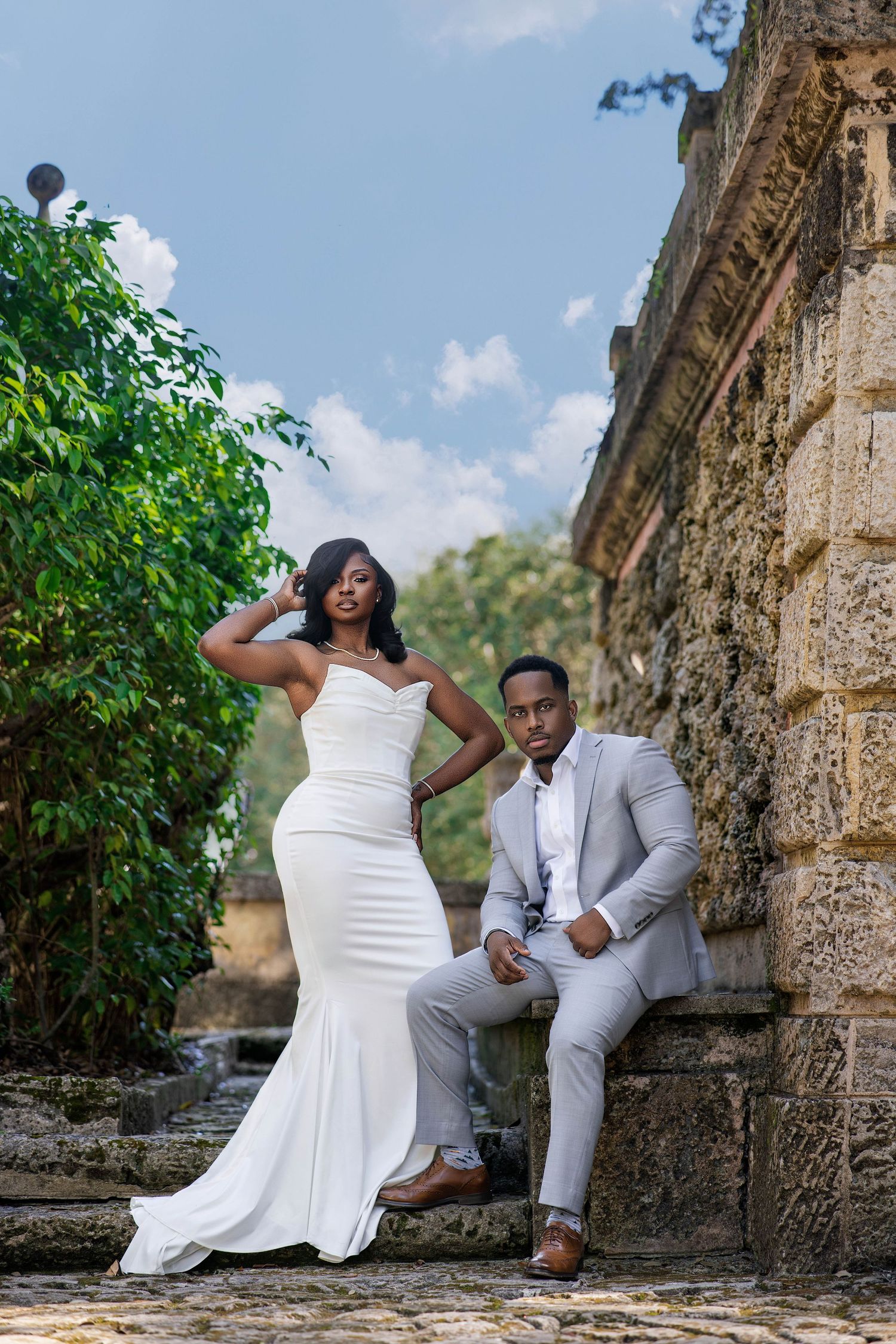 Elegant Black couple posing in wedding attire, bride in white mermaid gown and groom in gray suit, at stone ruins.