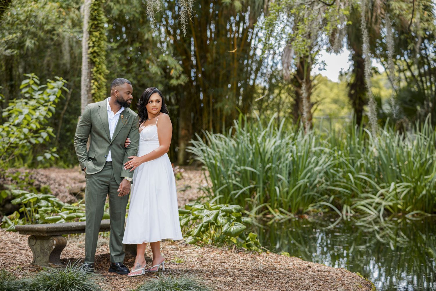 Interracial couple stands together near a serene pond surrounded by lush greenery.