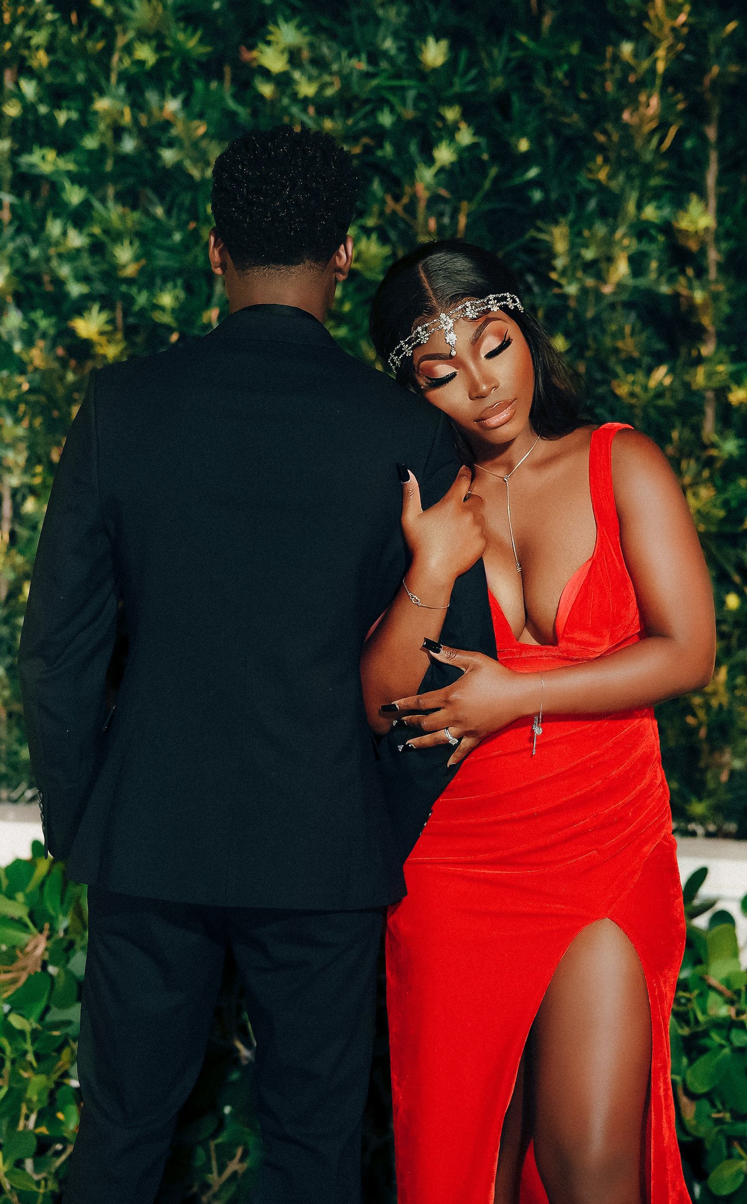 Couple in formal red and black attire share an intimate moment against a lush green hedge backdrop.