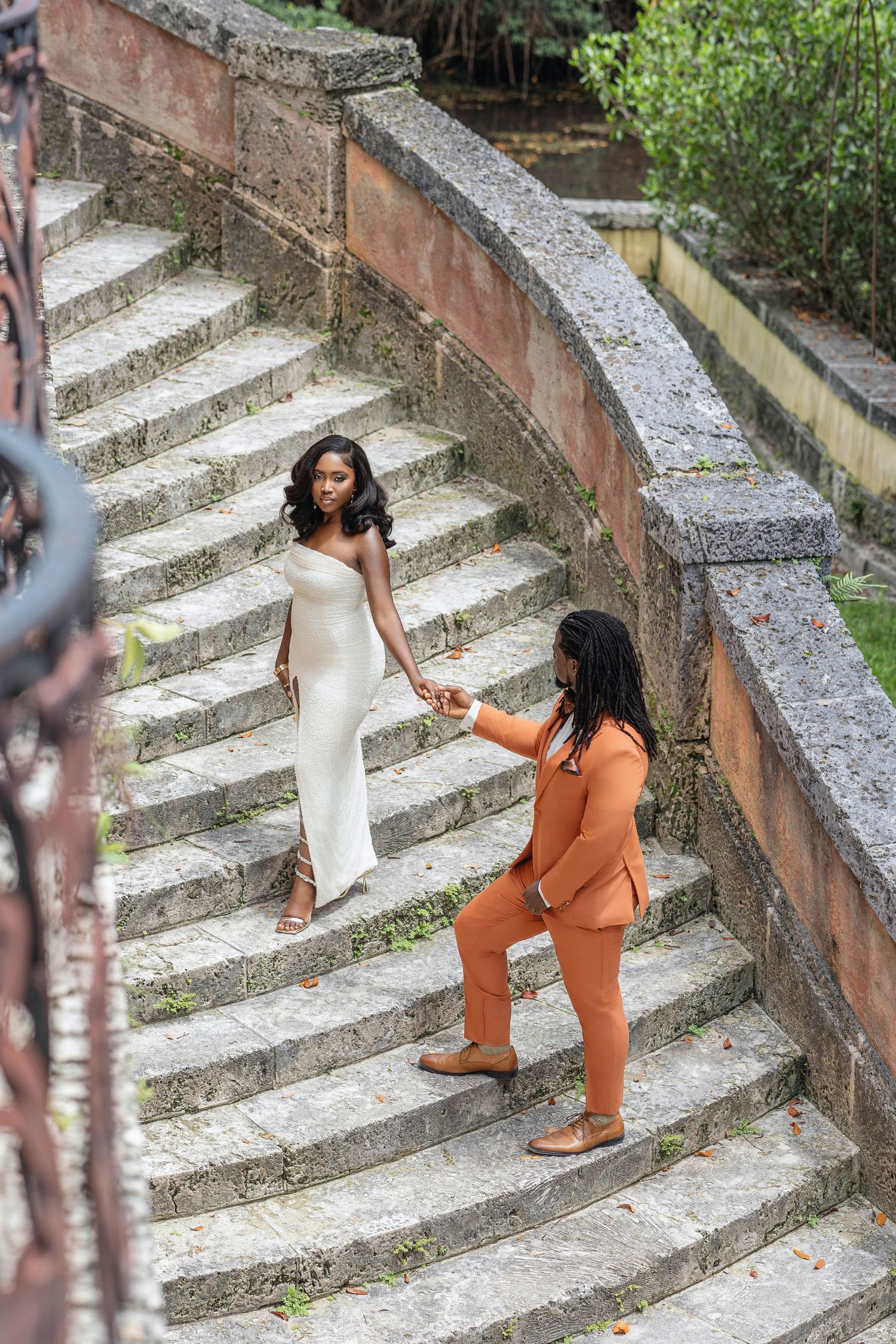 Groom in orange suit holds bride's hand on curved stone staircase in garden setting.