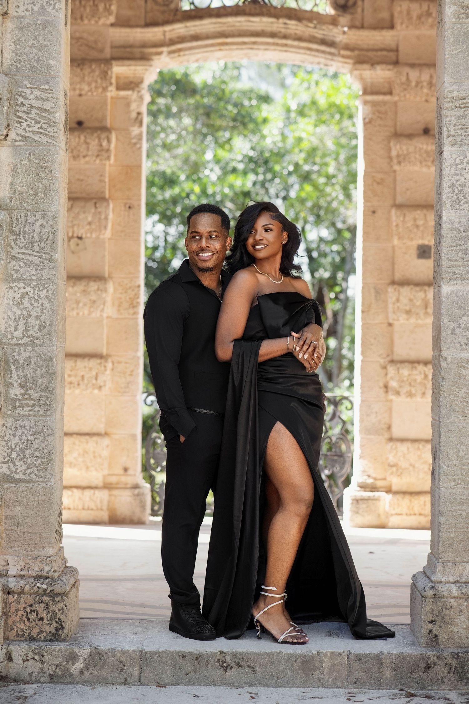 Black couple posing under stone archway, woman in elegant black gown with thigh slit and man in black attire.