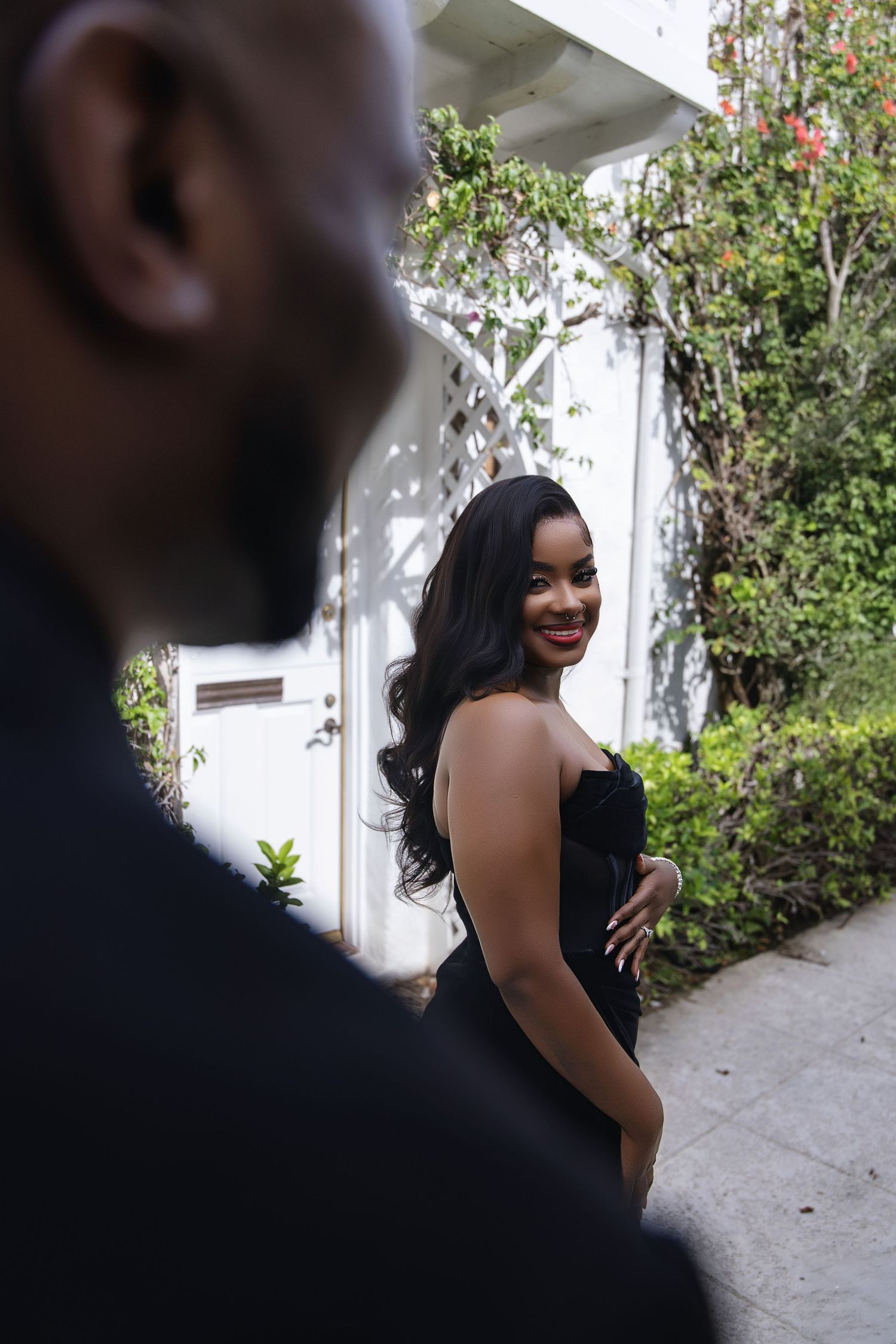Woman in elegant black strapless gown smiling over shoulder outdoors.